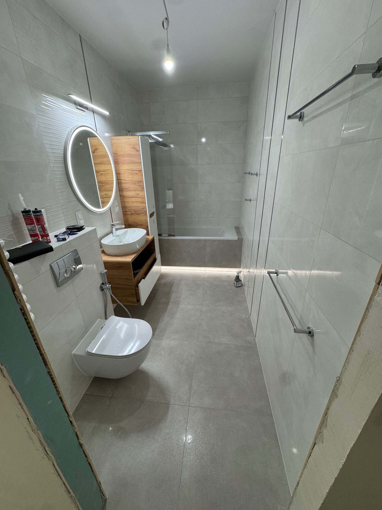 Nowoczesna łazienka z podświetlanym lustrem, drewnianą szafką pod umywalką i szarymi płytkami. Widoczna wanna za szklaną ścianką i grzejnik drabinkowy.