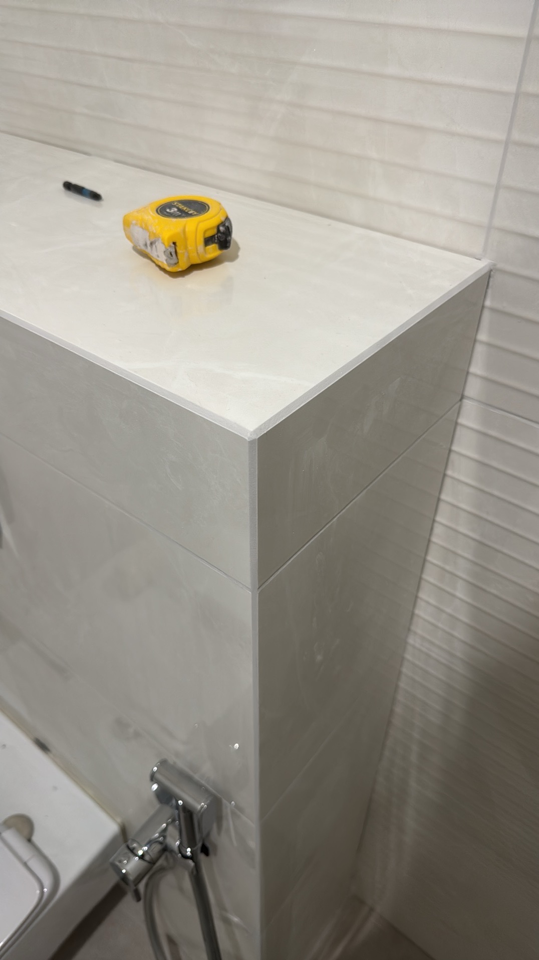 Wykończona obudowa WC z płytek w łazience, na blacie leży miara zwijana i ołówek. Na ścianie widoczne płytki z delikatnym wzorem. Widok z dołu.