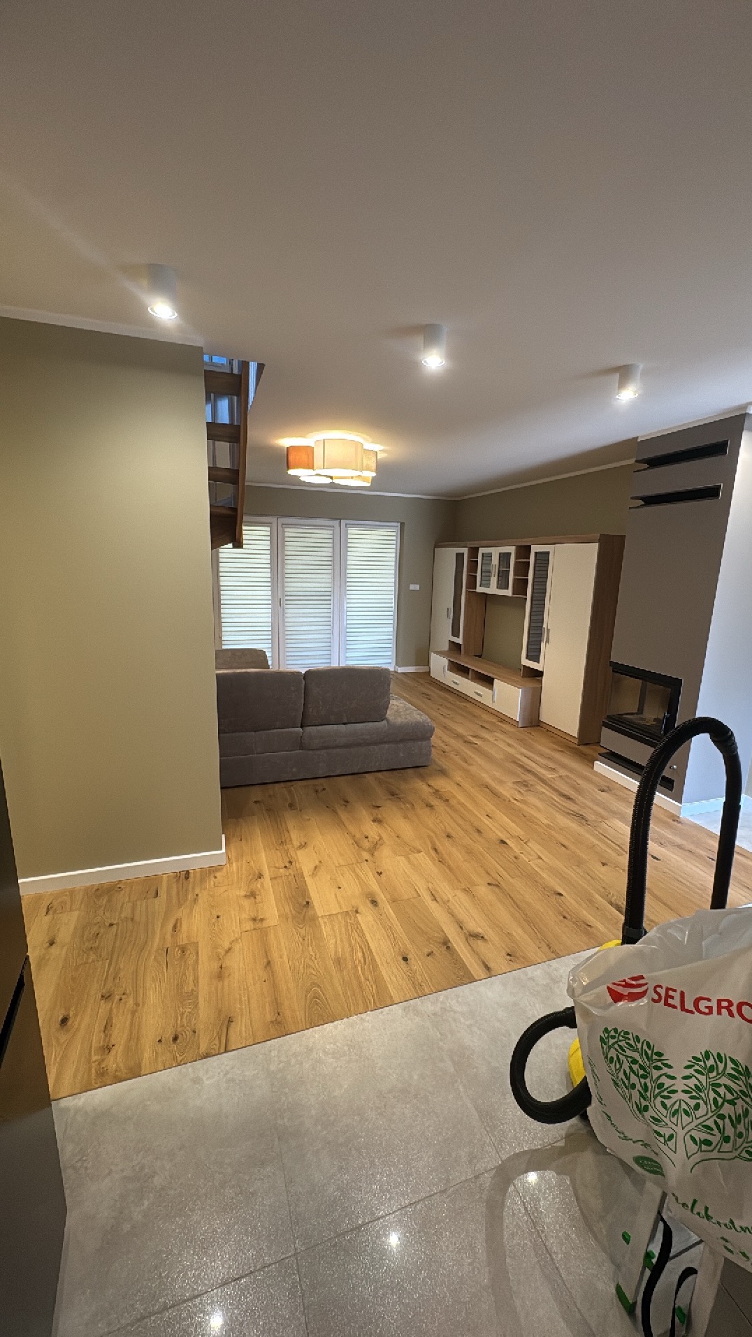 Jasne, przestronne wnętrze po remoncie: drewniana podłoga, szara sofa, białe meble. Widoczny fragment odkurzacza sugeruje trwające prace porządkowe.