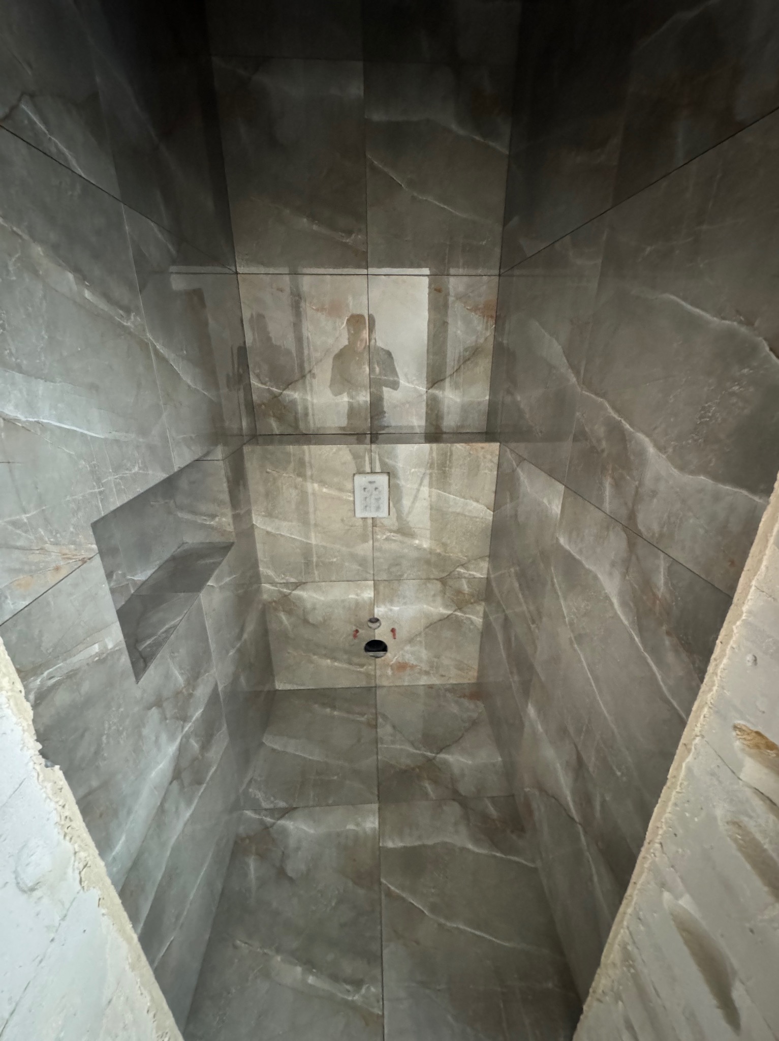 Wnętrze kabiny prysznicowej z szarymi płytkami imitującymi marmur, widok z dołu na sufit z odbiciem osoby robiącej zdjęcie. Widoczne niedokończone wykończenia.