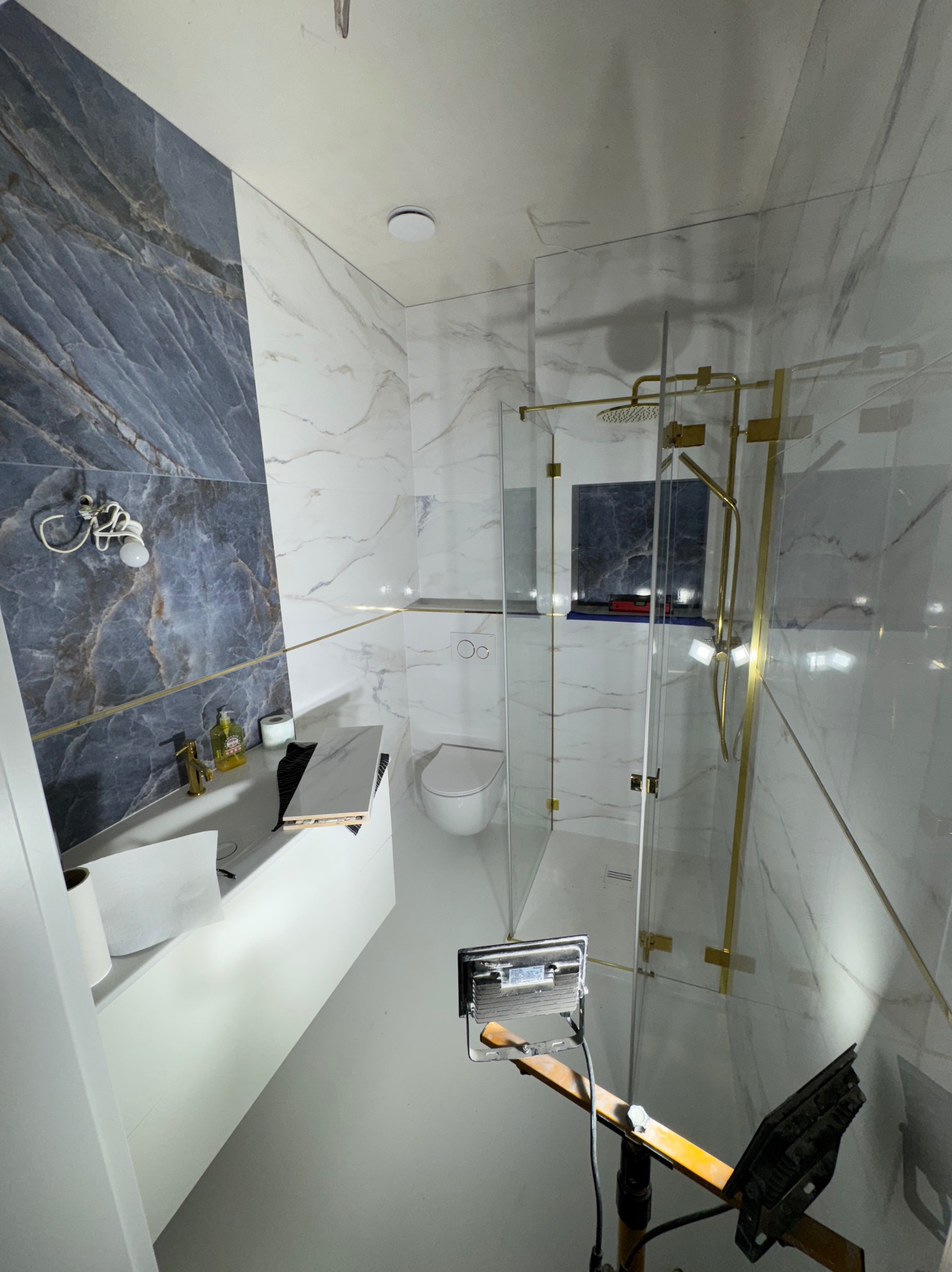 Nowoczesna łazienka z kabiną prysznicową typu walk-in, wykończona marmurowymi płytkami w odcieniach bieli i szarości. Widoczny fragment oświetlenia budowlanego.