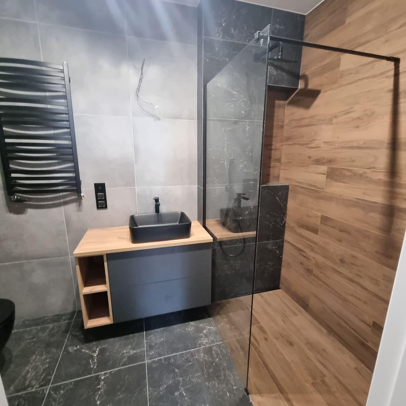 Nowoczesna łazienka z kabiną typu walk-in, umywalką nablatową i grzejnikiem drabinkowym. Szare płytki i drewnopodobne akcenty tworzą elegancki wystrój.