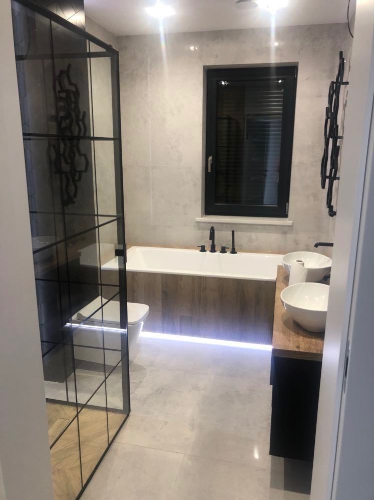 Nowoczesna łazienka z kabiną prysznicową typu walk-in z czarnymi szprosami, białą wanną z drewnianą obudową i podświetleniem LED oraz umywalkami nablatowymi.