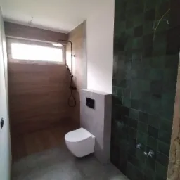 Wnętrze łazienki w trakcie wykańczania: widoczne zielone, błyszczące płytki na ścianie, drewnopodobne panele na ścianie prysznicowej, biała wisząca toaleta i szara posadzka.