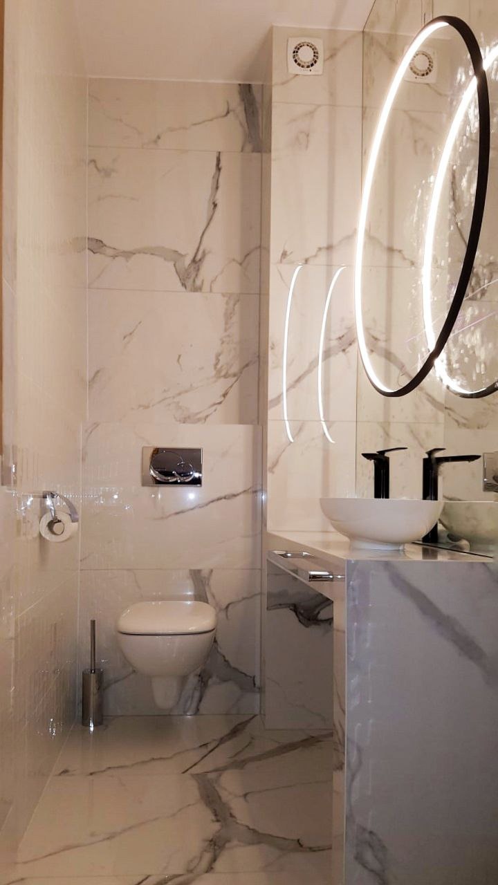 Nowoczesna łazienka wykończona płytkami imitującymi marmur, z podwieszaną toaletą, umywalką nablatową i okrągłym lustrem z podświetleniem LED.