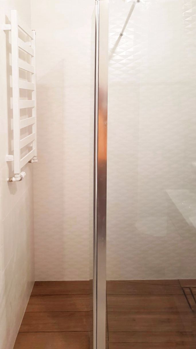 Fragment łazienki z kabiną prysznicową z chromowanymi profilami, grzejnikiem drabinkowym na ścianie i drewnopodobną podłogą z płytek.
