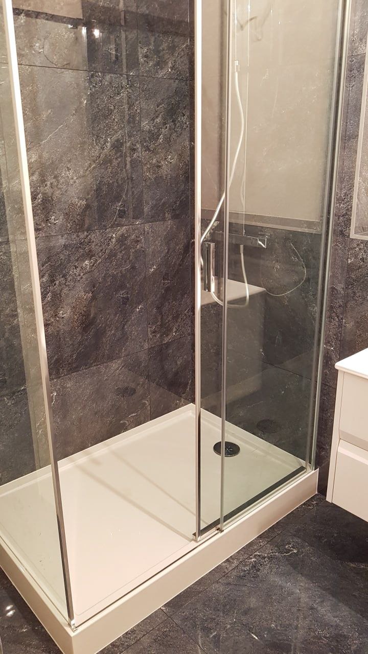 Kabina prysznicowa z przesuwnymi drzwiami, biały brodzik i ciemnoszara okładzina z płytek imitujących kamień na ścianach i podłodze. Widoczna bateria prysznicowa i biała szafka pod umywalkę.