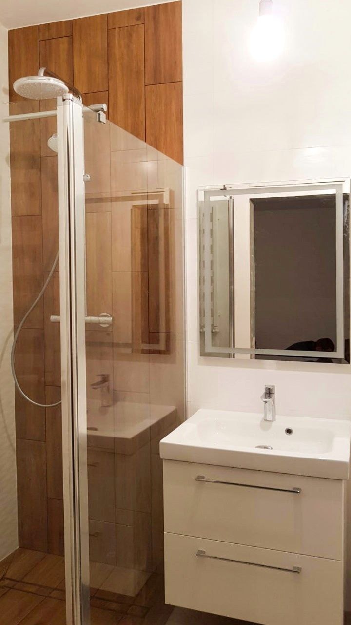 Nowoczesna łazienka z kabiną prysznicową z drewnopodobnymi płytkami, białą umywalką z szafką i podświetlanym lustrem.