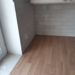 Tomasz Jankosz - Fragment kuchni po remoncie: ułożone płytki imitujące cegłę w odcieniach szarości nad blatem z paneli drewnopodobnych, widoczny fragment okna i szafki kuchennej z drewnianymi gałkami.