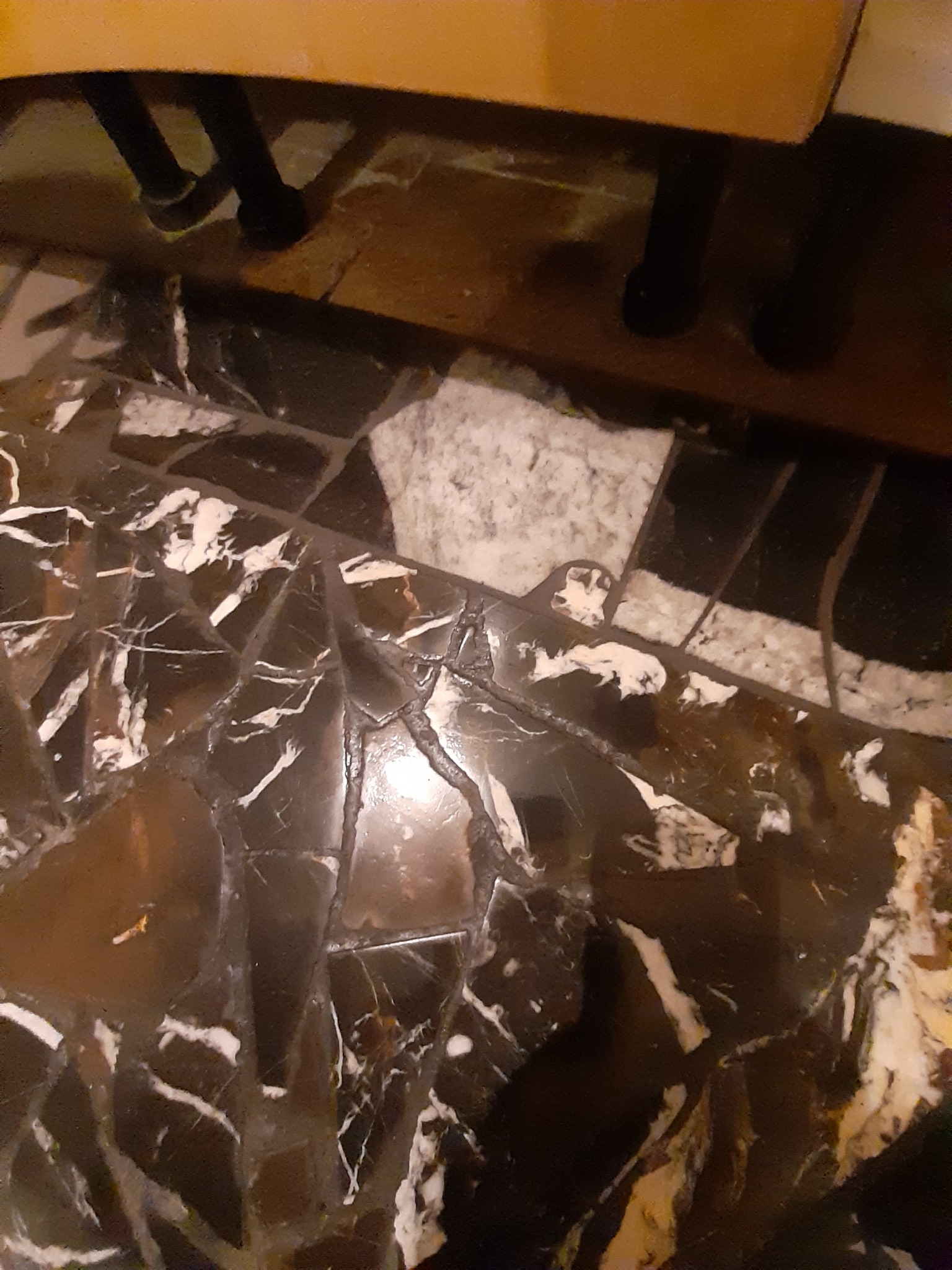 Fragment podłogi z czarno-białych płytek imitujących marmur, widoczne nogi krzesła w tle.