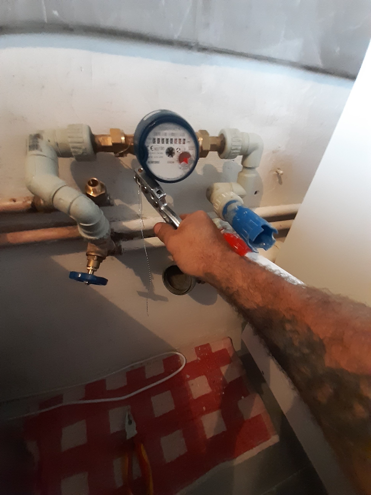 Instalacja wodomierza z rurami PCV i zaworem, przy użyciu klucza nastawnego, na tle ściany i podłogi wyłożonej płytkami z wzorem.