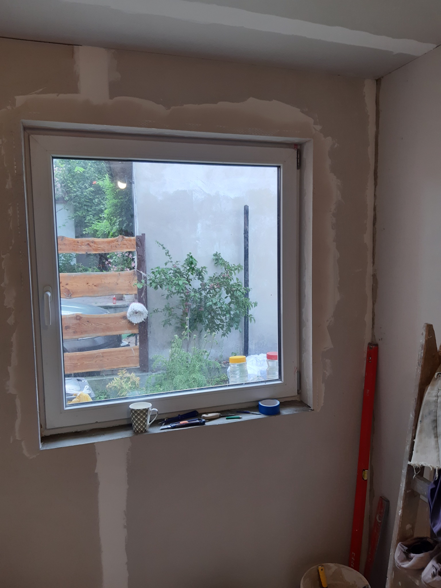 Przygotowanie ściany wokół okna do malowania, widoczne ślady gładzi szpachlowej, poziomica, drabina, kubek i narzędzia na parapecie.