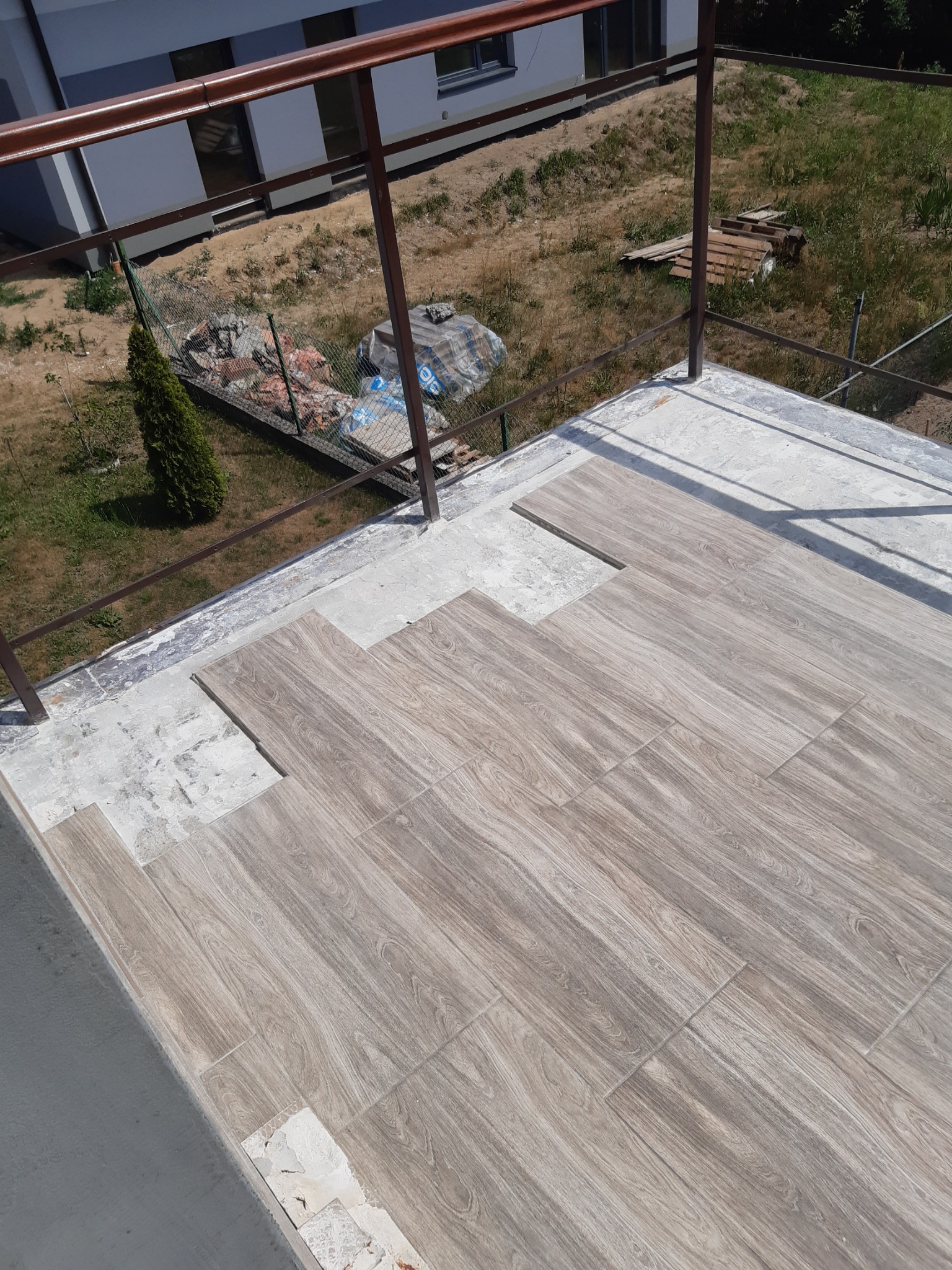 Układanie płytek imitujących drewno na balkonie, widoczny etap prac, niezakończona powierzchnia, szara balustrada w tle.