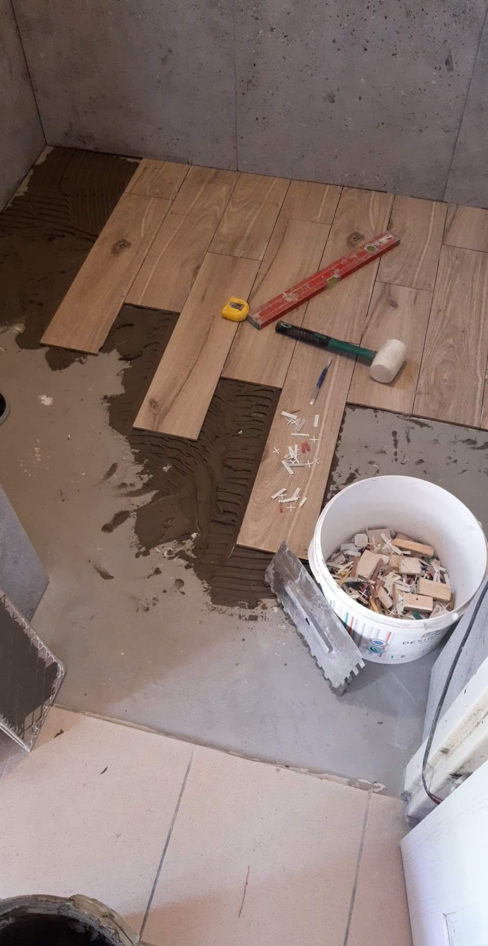 Proces układania płytek imitujących drewno w pomieszczeniu, widoczny klej, poziomica, gumowy młotek i wiadro z elementami dystansowymi.