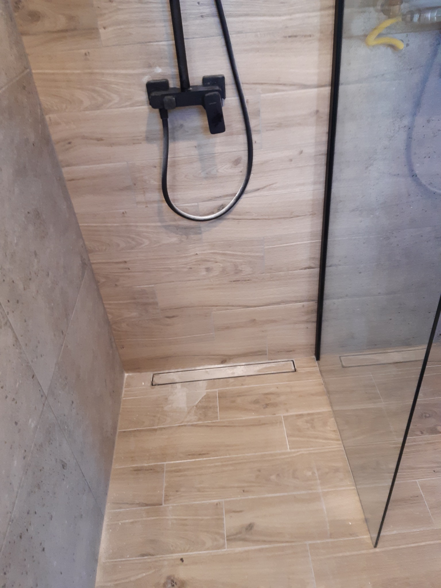 Wykończona kabina prysznicowa z odpływem liniowym, wyłożona płytkami imitującymi drewno, czarna armatura natryskowa.