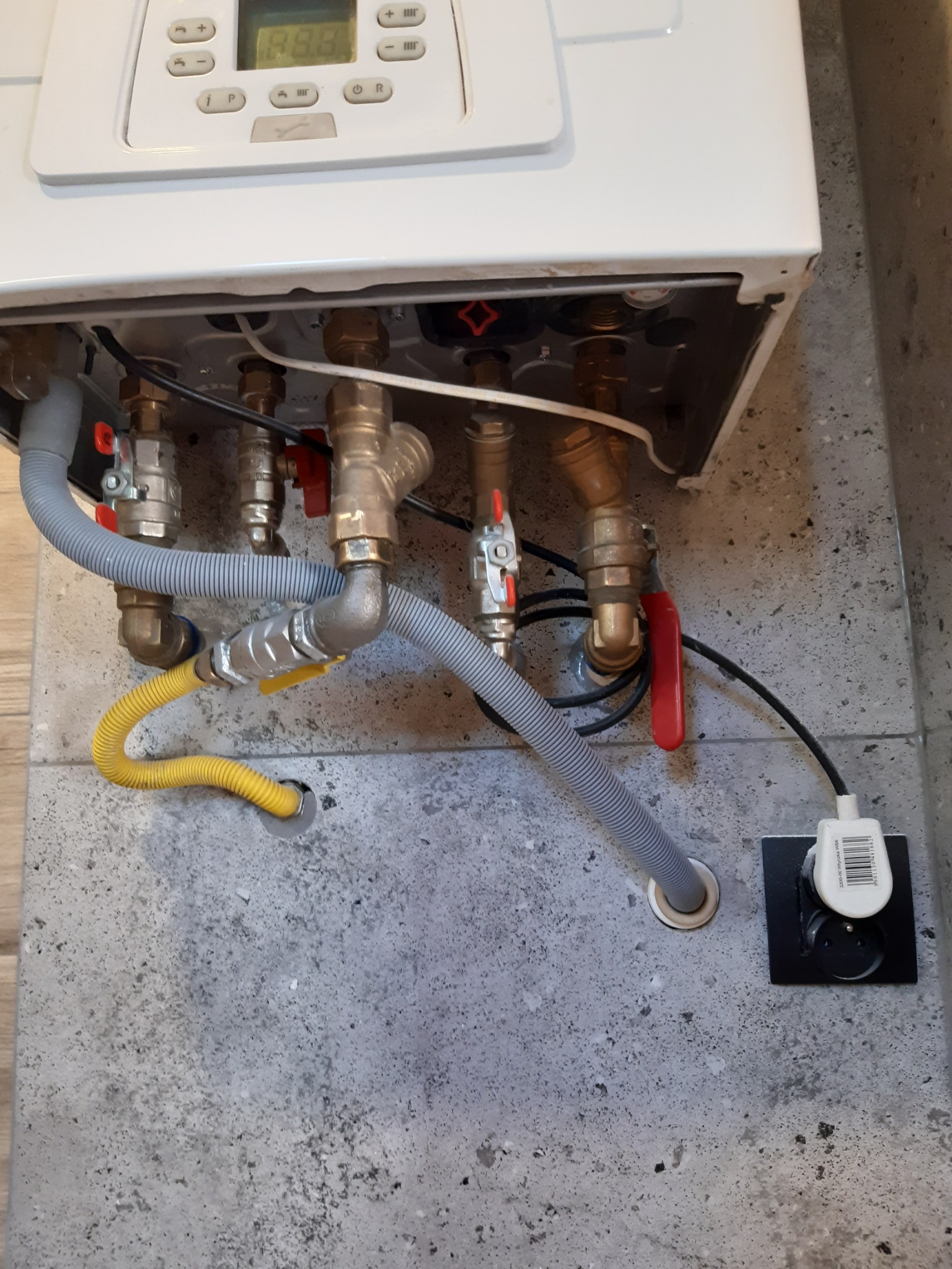 Podłączenie gazowego pieca centralnego ogrzewania do instalacji: widoczne rury, zawory, przewód gazowy w żółtej osłonie oraz gniazdko elektryczne w ścianie o szarej, betonowej fakturze.