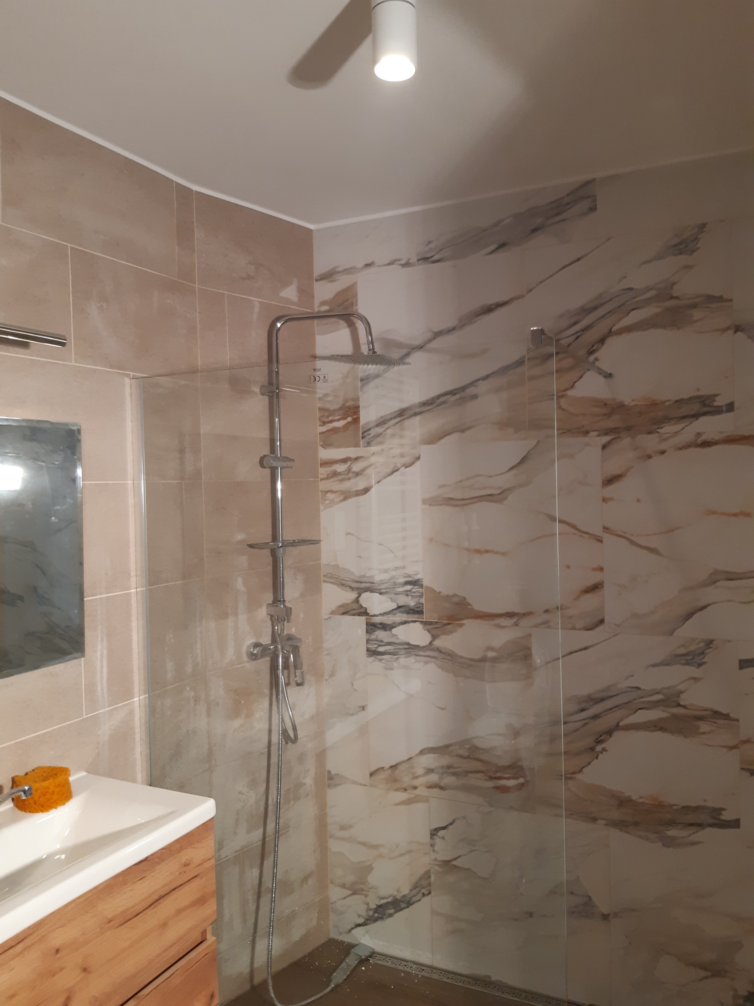 Nowoczesna kabina prysznicowa z przeszkloną ścianką, wykończona płytkami imitującymi marmur, widoczny drewniany blat umywalki i chromowana bateria.