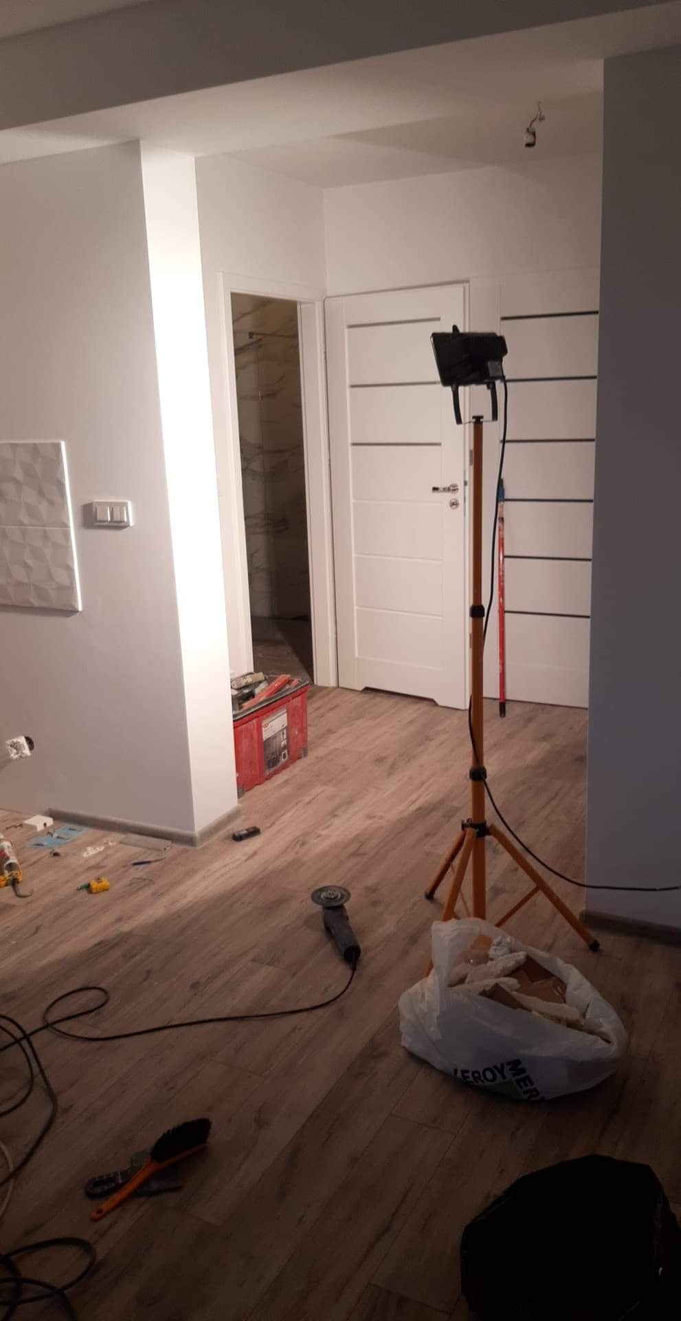Wnętrze w trakcie prac wykończeniowych: widoczna lampa budowlana na statywie, szlifierka kątowa, torba z odpadami budowlanymi oraz świeżo położona podłoga imitująca drewno i białe ściany...