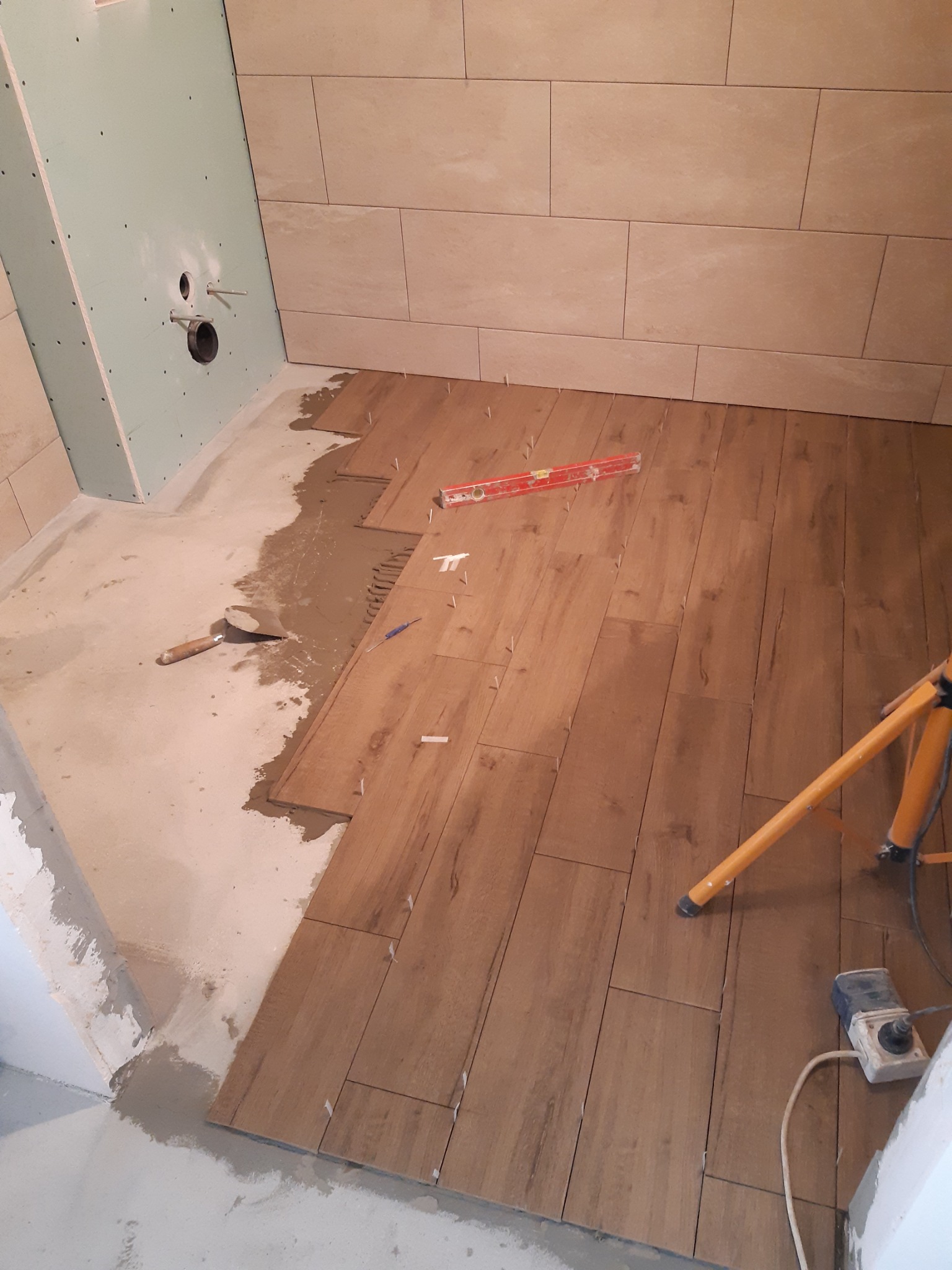 Układanie płytek imitujących drewno na podłodze w łazience, widoczny poziomica, szpachelka i lampa budowlana.