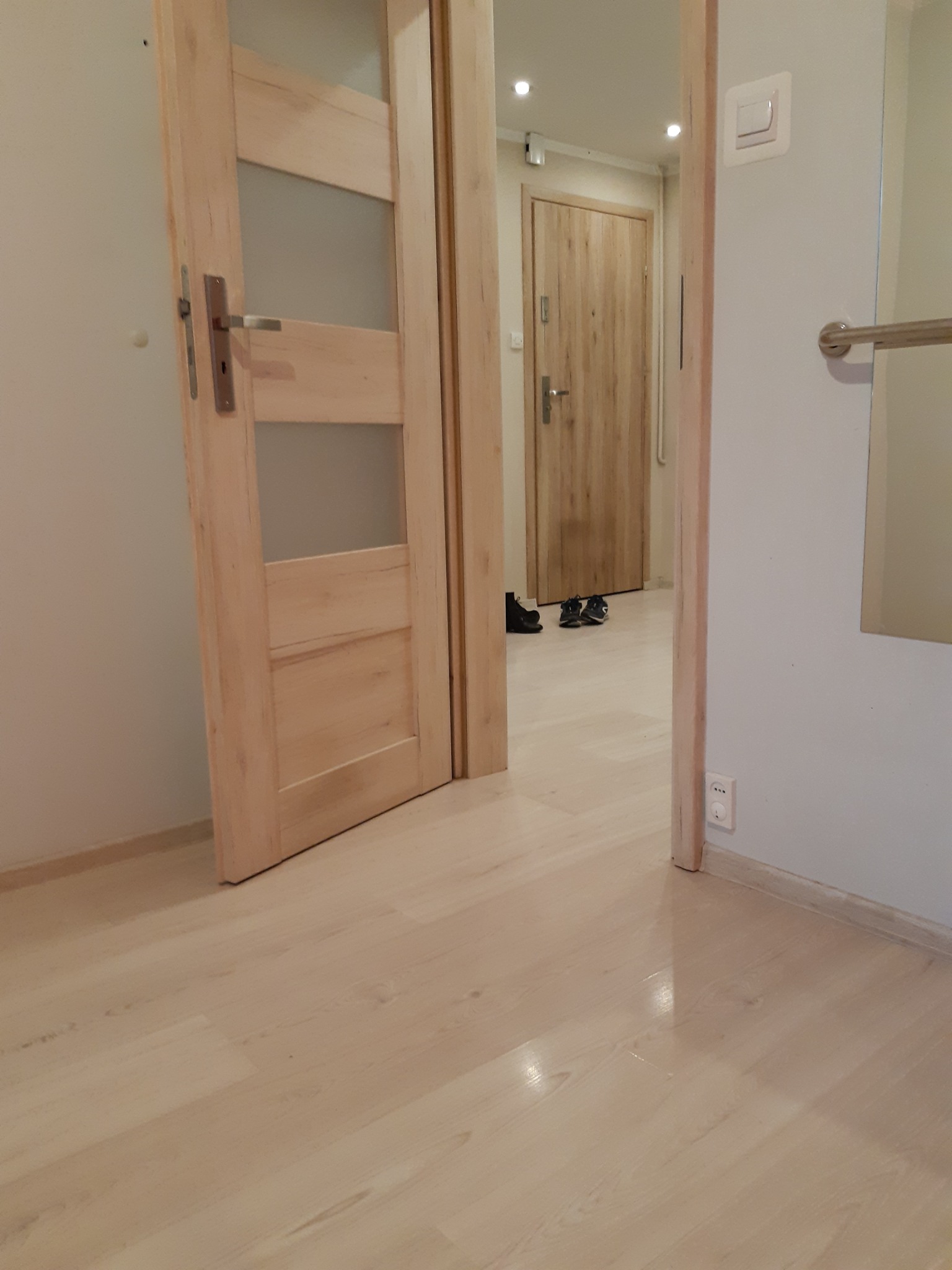 Otwarta drewniana wewnętrzna drzwi z trzema szybkami w jasnym korytarzu, widoczna druga, zamknięta drewniana drzwi w tle, na podłodze leżą buty.