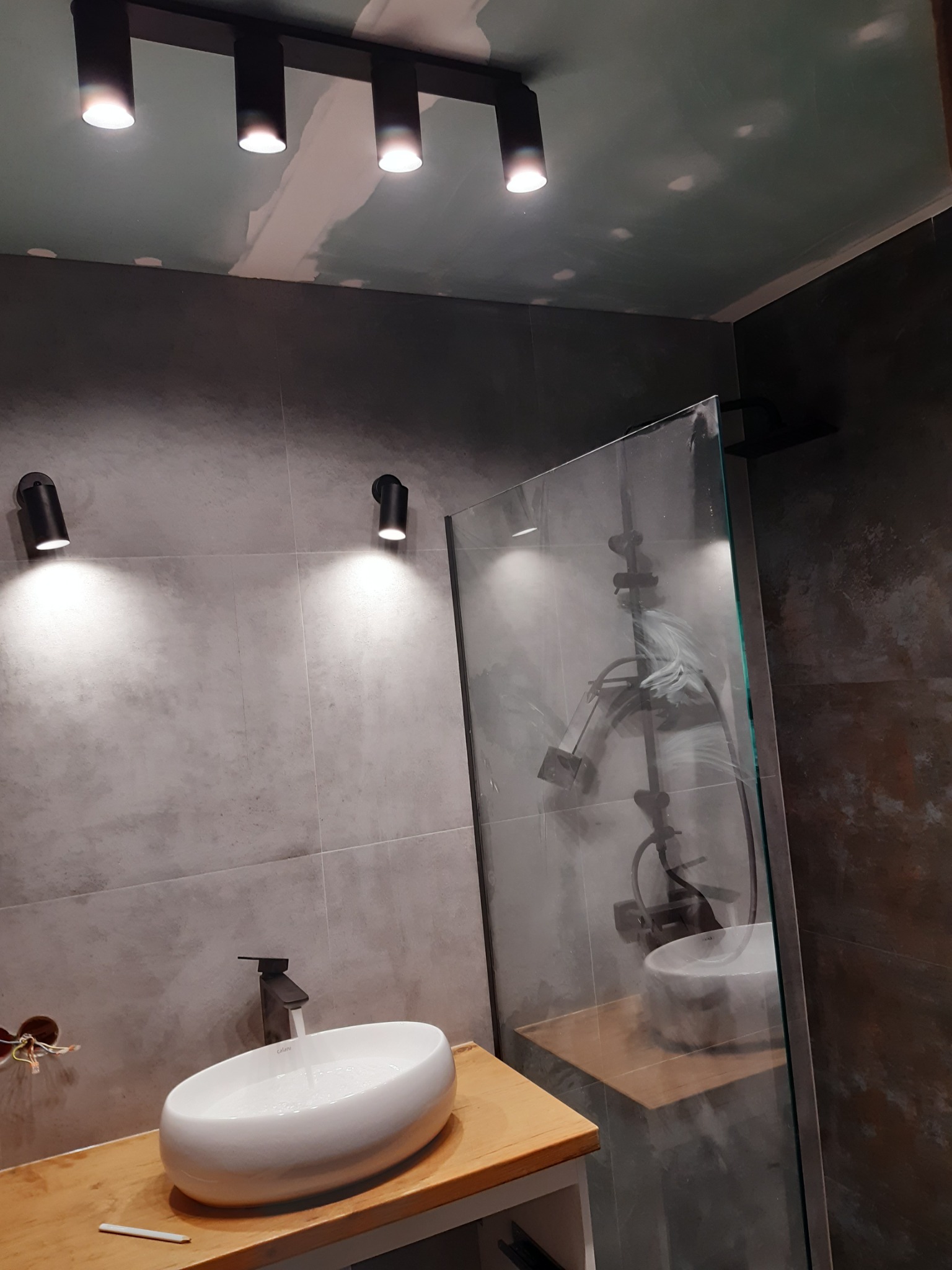 Szara łazienka z owalną umywalką nablatową, baterią w kolorze czarnym, kabiną prysznicową ze szklaną ścianką, oświetleniem punktowym na suficie i ścianie, widoczne ślady po szpachlowaniu na suficie.