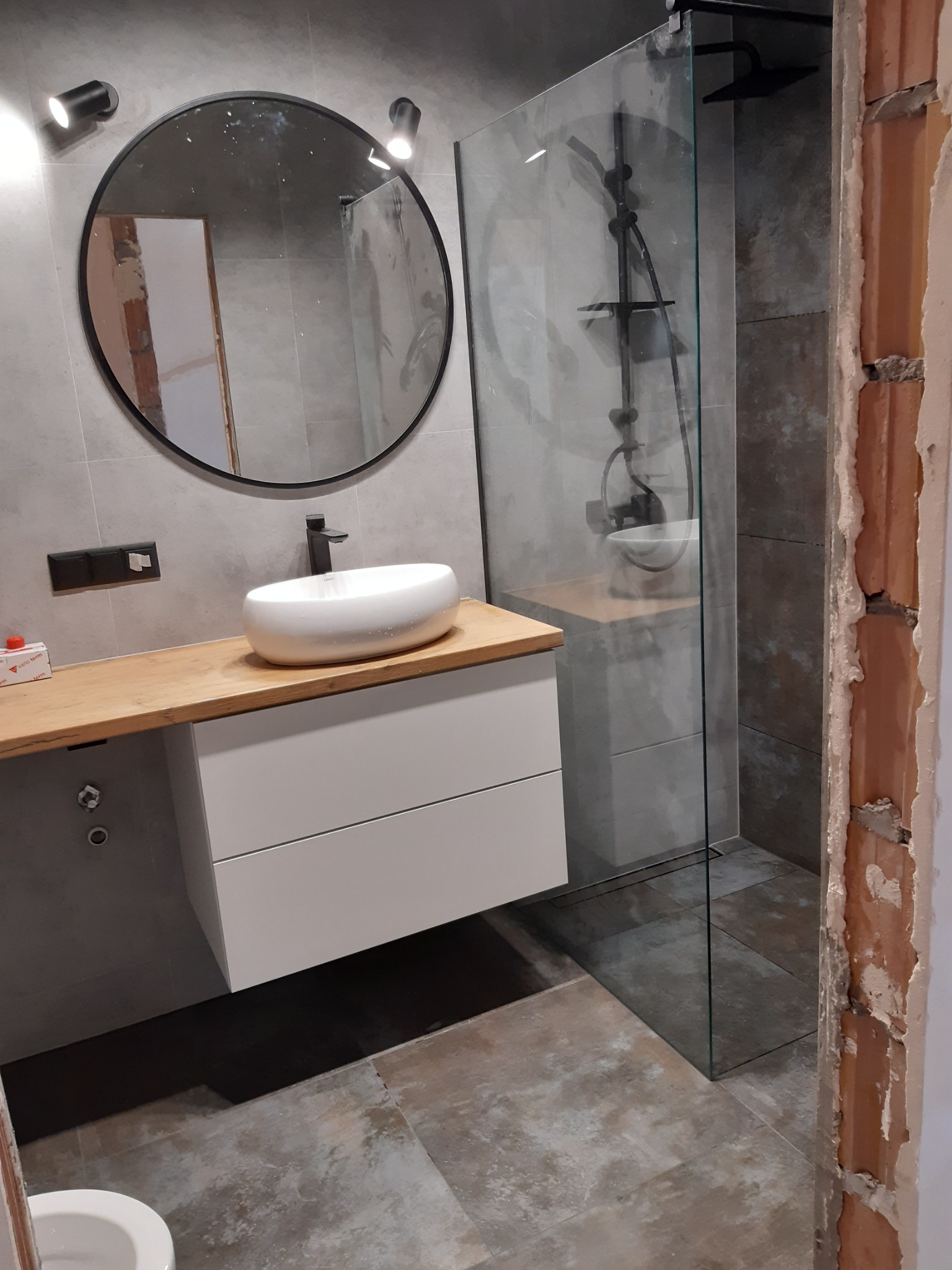 Nowoczesna łazienka w trakcie wykończenia: szare płytki, okrągłe lustro z czarną ramą nad białą umywalką na drewnianym blacie, kabina prysznicowa ze szklaną ścianą i czarną armaturą, widoczne...