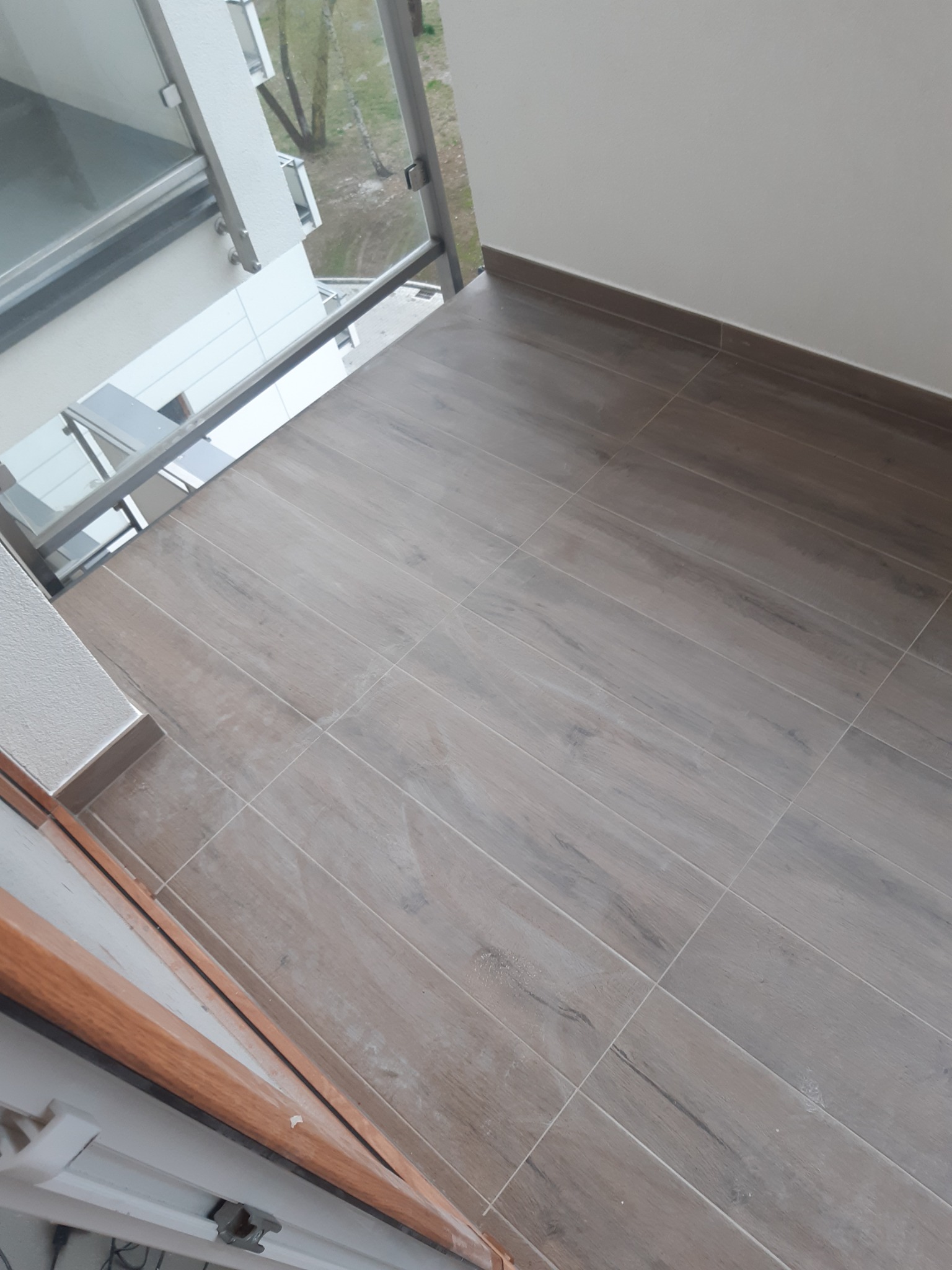 Balkon wyłożony płytkami imitującymi drewno, z widoczną szklaną balustradą i fragmentem ramy okiennej