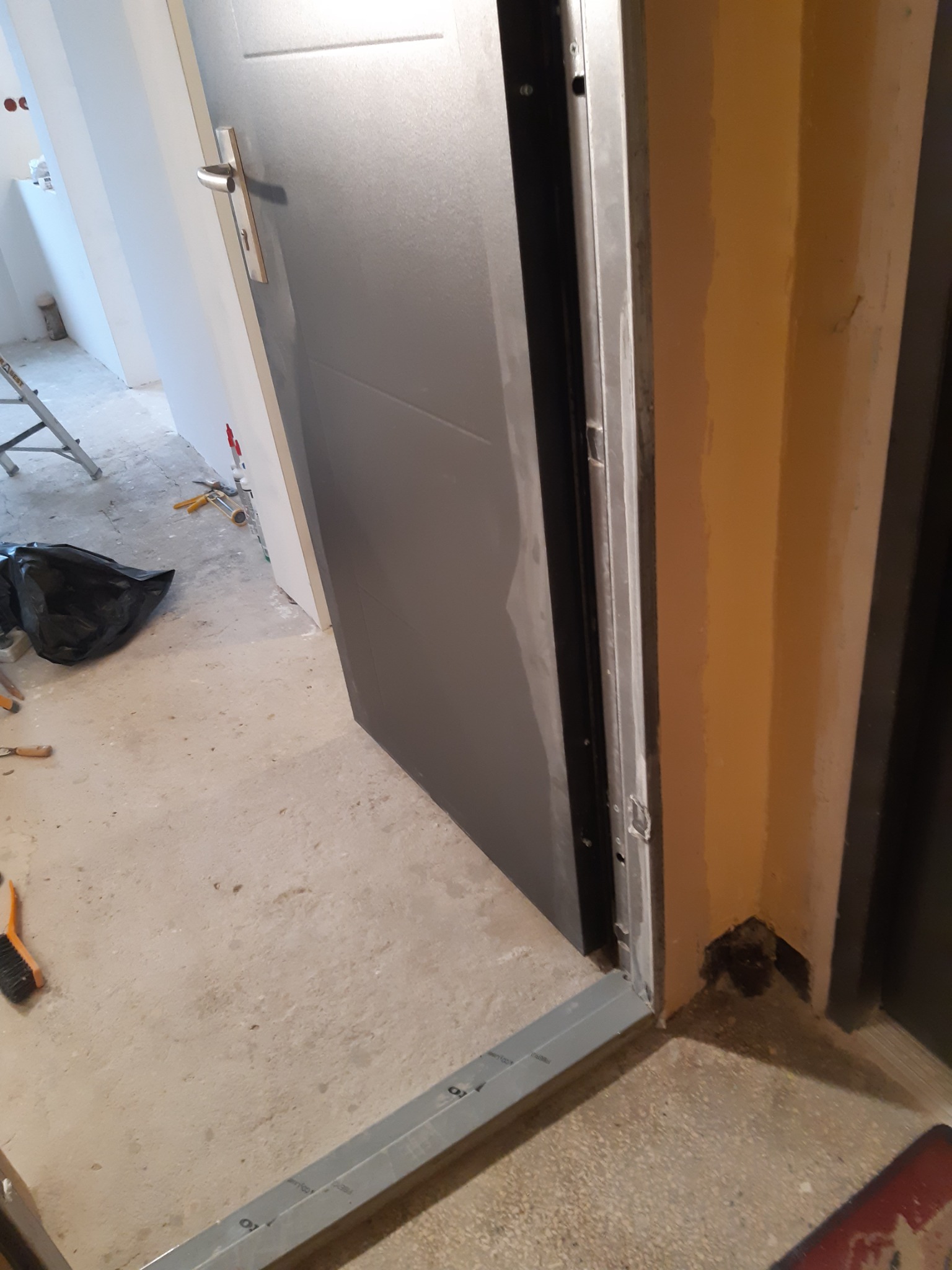 Szare drzwi wejściowe w trakcie montażu, widoczna metalowa ościeżnica, surowa podłoga i narzędzia w tle.