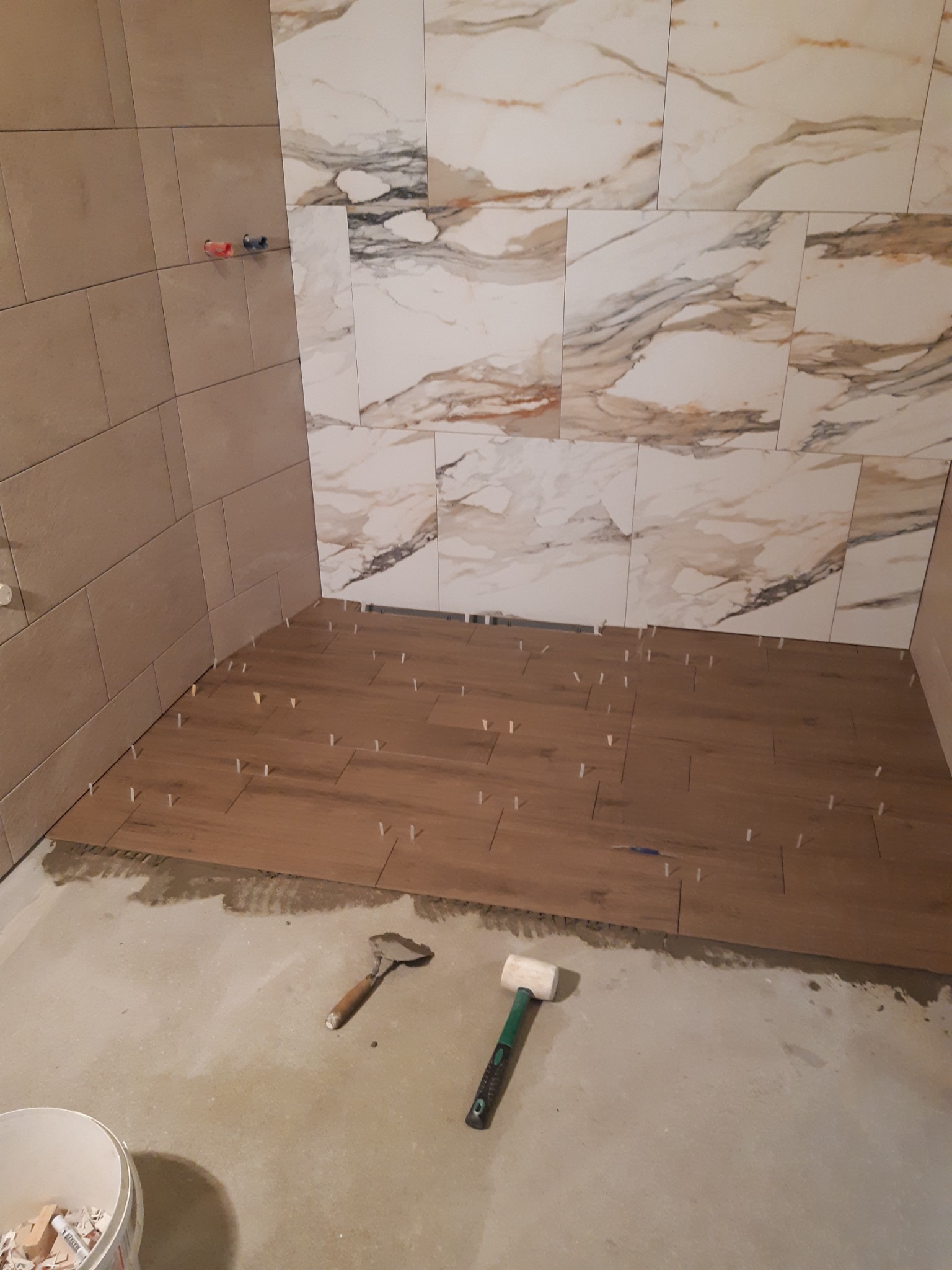 Proces układania płytek podłogowych imitujących drewno z użyciem krzyżyków dystansowych; na ścianie płytki imitujące marmur, widoczna kielnia i gumowy młotek.