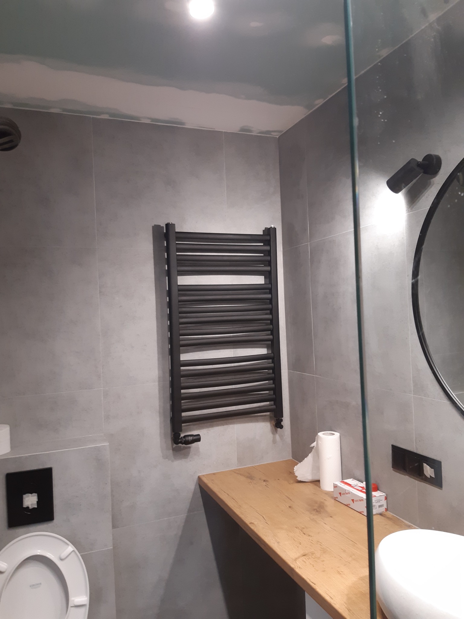 Szara łazienka w trakcie remontu z zamontowanym czarnym grzejnikiem drabinkowym, drewnianym blatem, białą umywalką i toaletą.