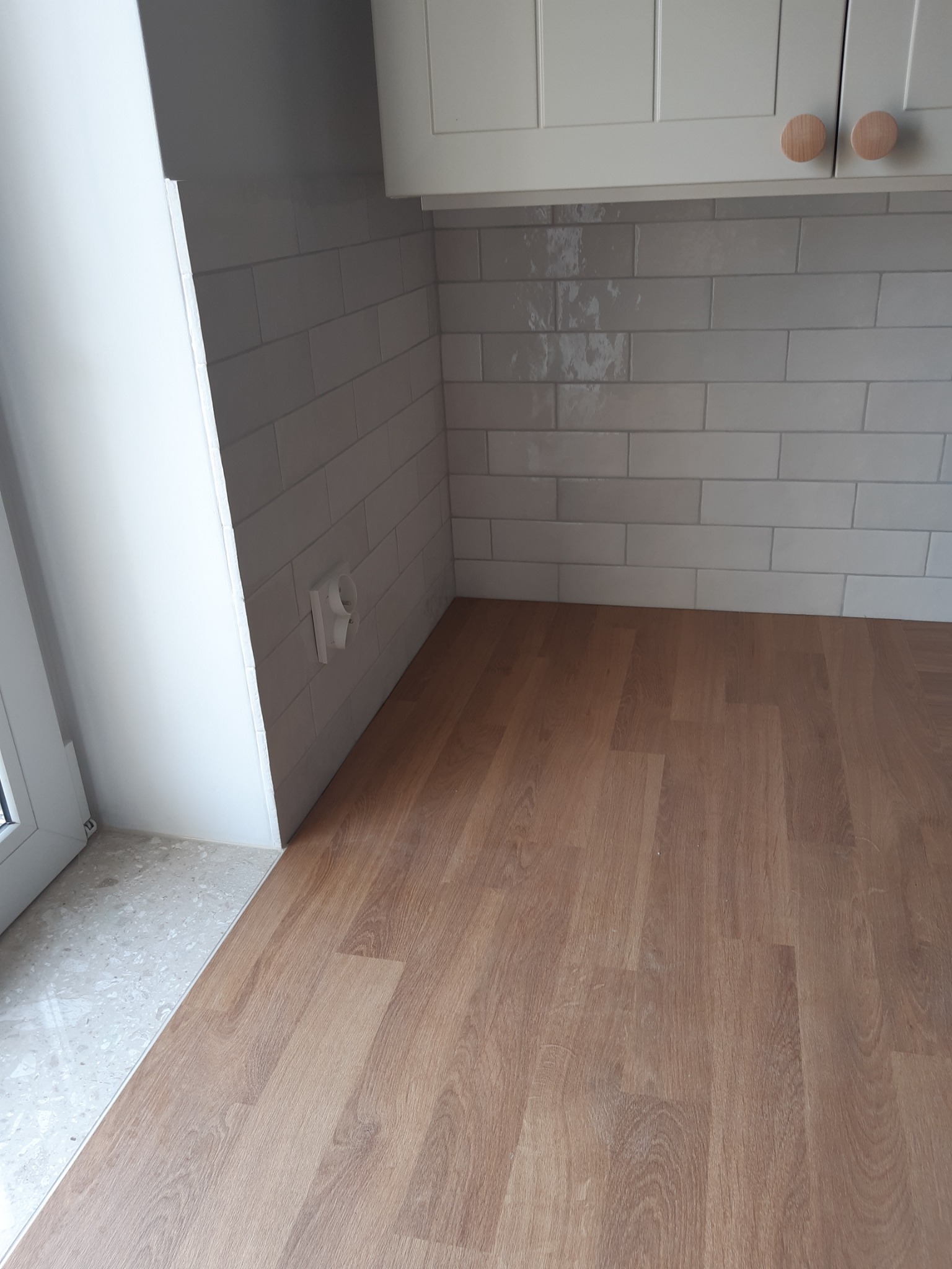 Fragment kuchni po remoncie: ułożone płytki imitujące cegłę w odcieniach szarości nad blatem z paneli drewnopodobnych, widoczny fragment okna i szafki kuchennej z drewnianymi gałkami.