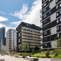 KONCEPT Nieruchomości - Nowoczesne budynki mieszkalne z balkonami, widoczne z poziomu chodnika z zielenią miejską i ławkami; w tle wieżowiec w budowie z dźwigiem.