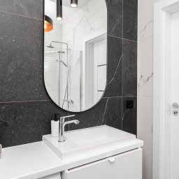 KONCEPT Nieruchomości - Nowoczesna łazienka z czarną mozaikową ścianą, owalnym lustrem, białą umywalką nablatową, baterią chromowaną i stosem szarych ręczników.