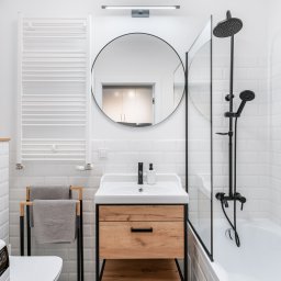 KONCEPT Nieruchomości - Jasna łazienka z białymi płytkami, okrągłym lustrem, drewnianą szafką pod umywalką i czarną armaturą.