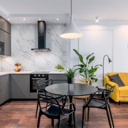 KONCEPT Nieruchomości - Nowoczesna kuchnia z szarymi szafkami, marmurowym backsplashiem, czarnym okrągłym stołem otoczonym czarnymi krzesłami, oraz żółtą kanapą z portretem Salvadora Dali w tle.