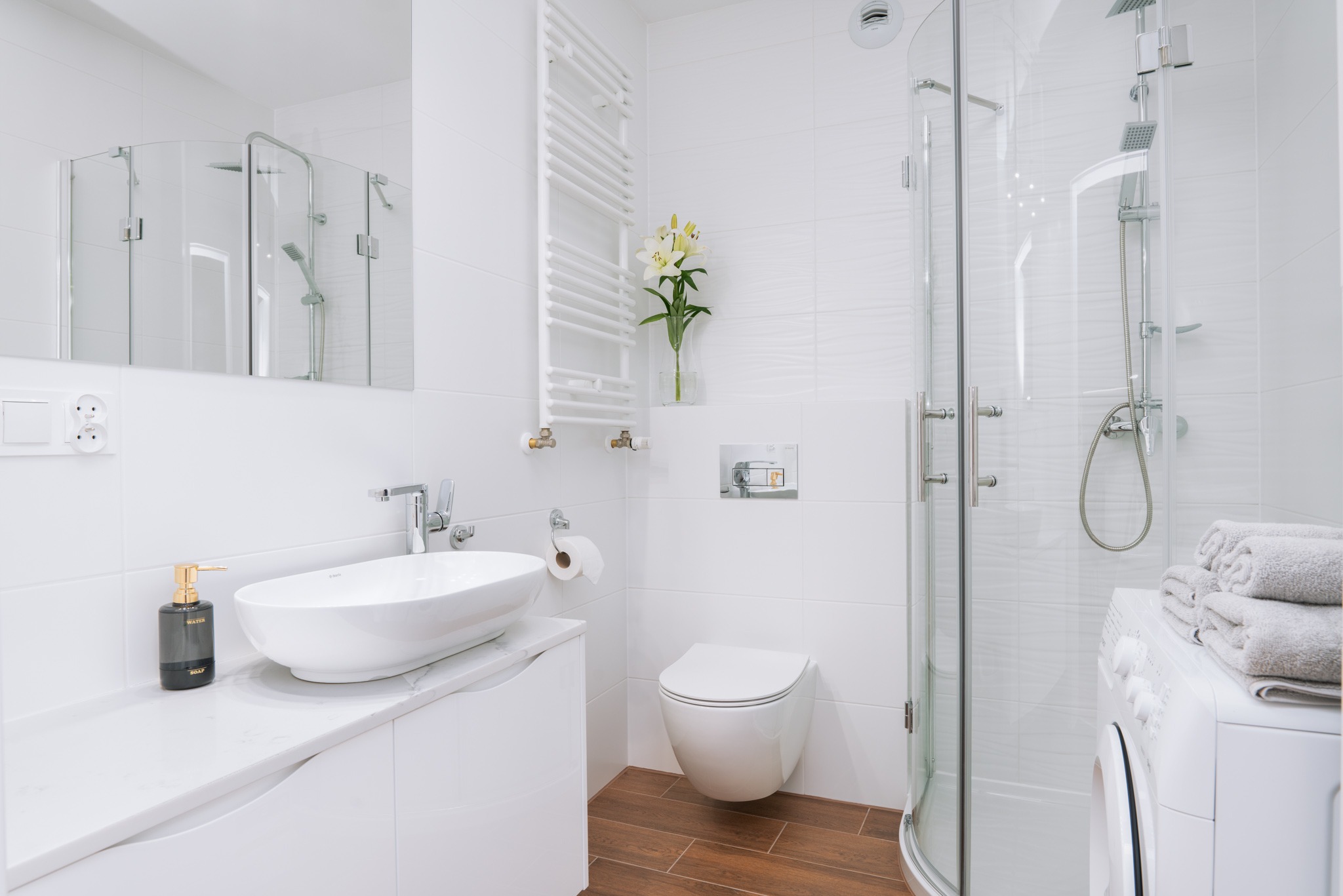 Jasna, nowoczesna łazienka z drewnopodobną podłogą, owalną umywalką na szafce, wiszącą toaletą, kabiną prysznicową i pralką z ręcznikami.