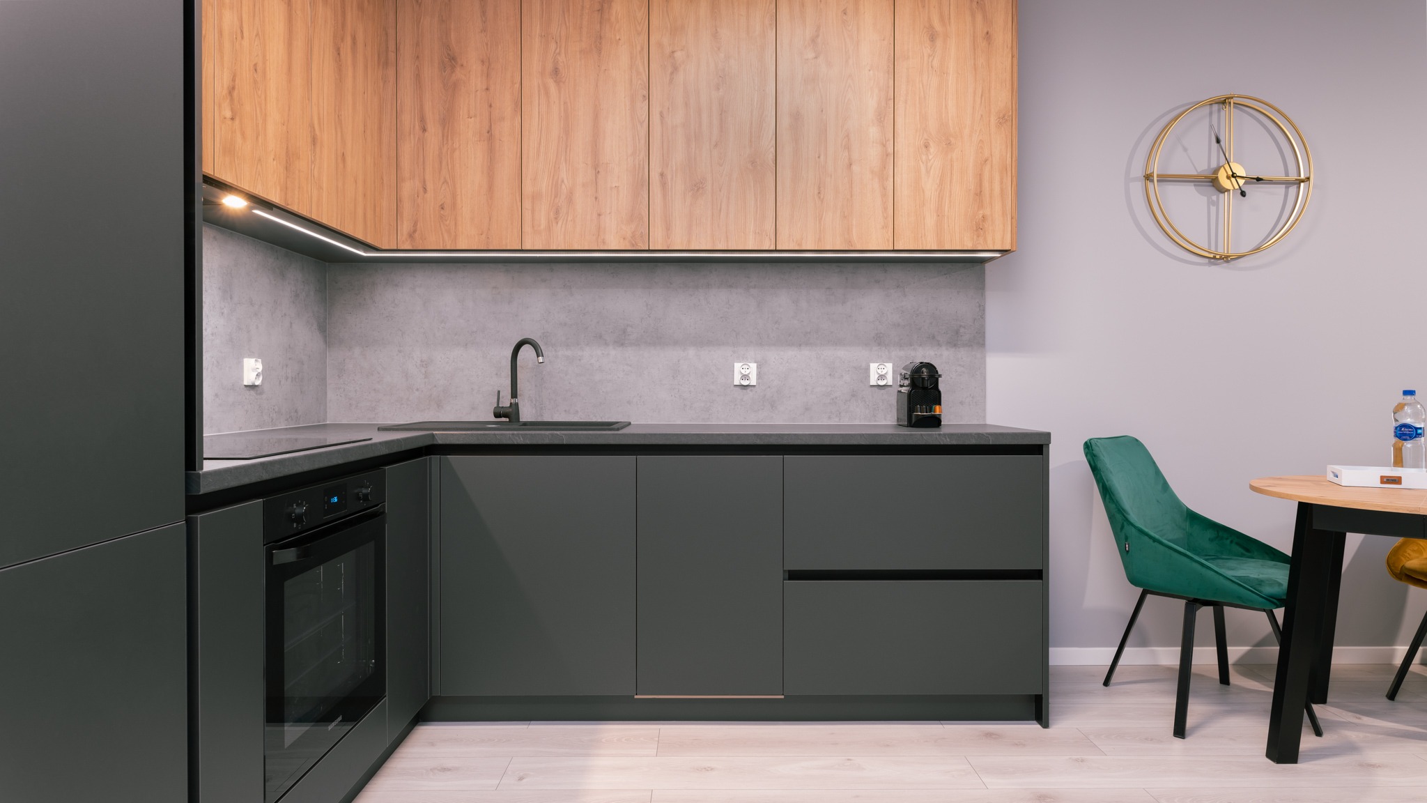 Nowoczesna kuchnia z szarymi szafkami, drewnianymi górnymi szafkami, kamiennym blatem i złotym zegarem na szarej ścianie, uzupełniona zielonym krzesłem.