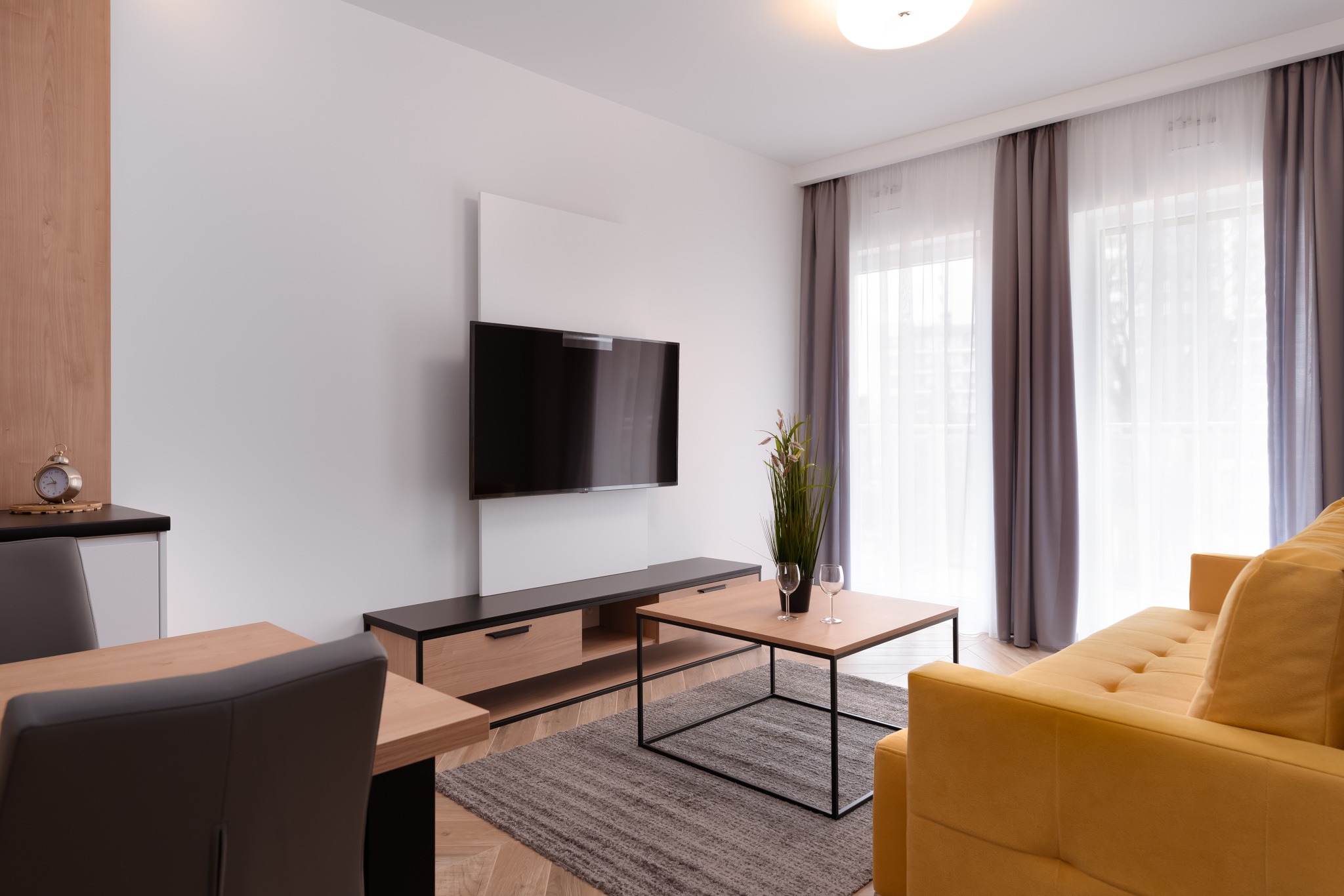Jasny salon z żółtą sofą, telewizorem na ścianie, drewnianym stolikiem kawowym i dywanem w odcieniach szarości, minimalistyczny wystrój, duże okno z zasłonami.