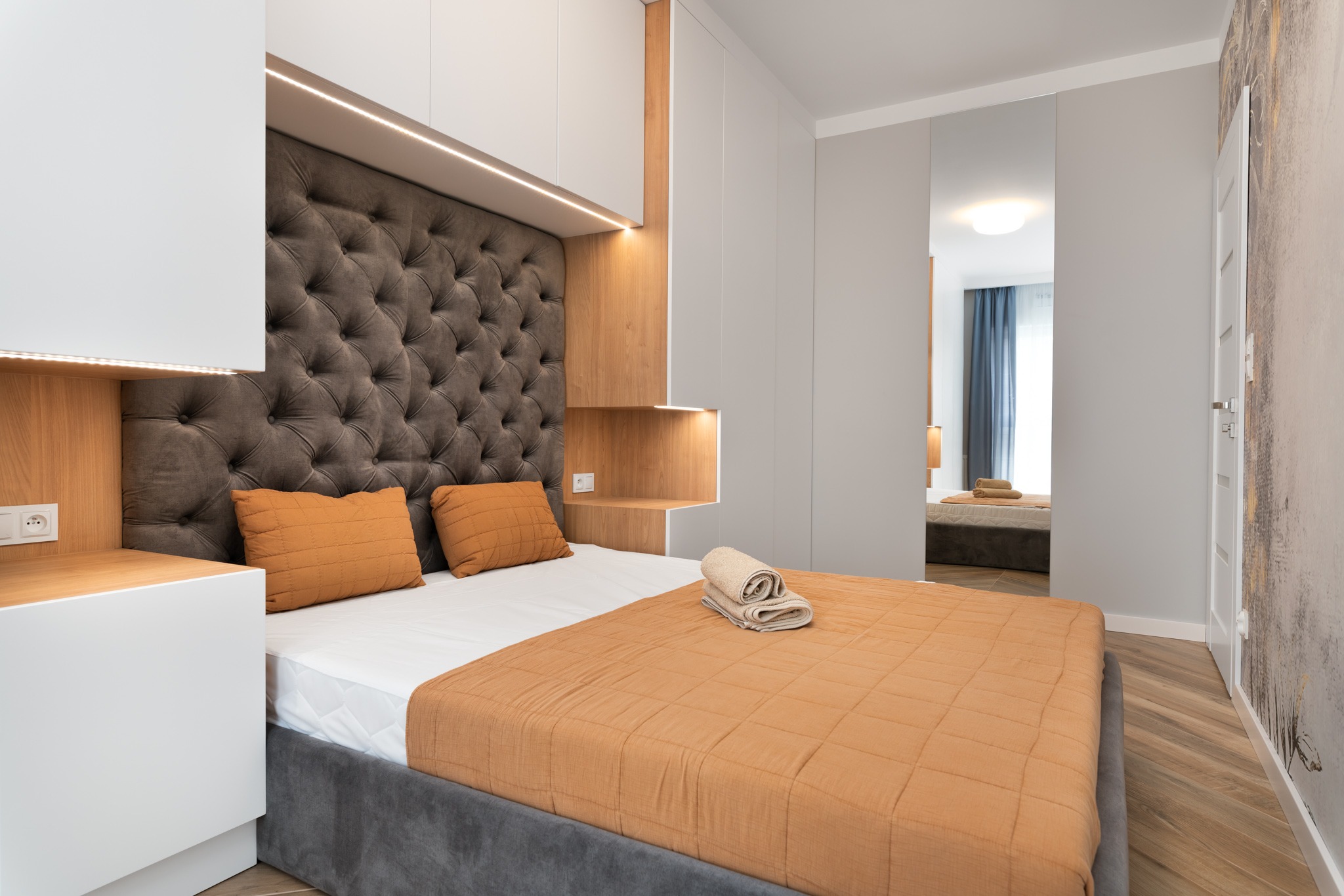 Nowoczesna sypialnia z szarą tapicerowaną ścianą z guzikami, pomarańczowymi poduszkami i narzutą, białymi szafkami i drewnianymi elementami, oświetleniem LED oraz lustrzanymi drzwiami przesuwnymi...