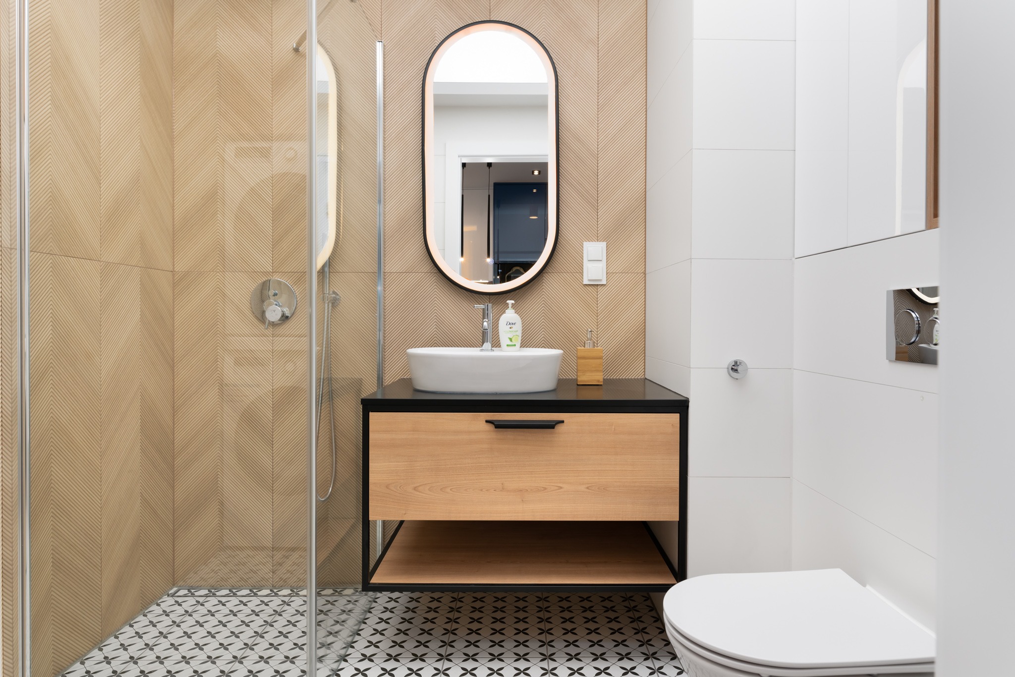 Nowoczesna łazienka z kabiną prysznicową, umywalką nablatową na szafce z drewnianym frontem, podświetlanym lustrem i wzorzystą podłogą.