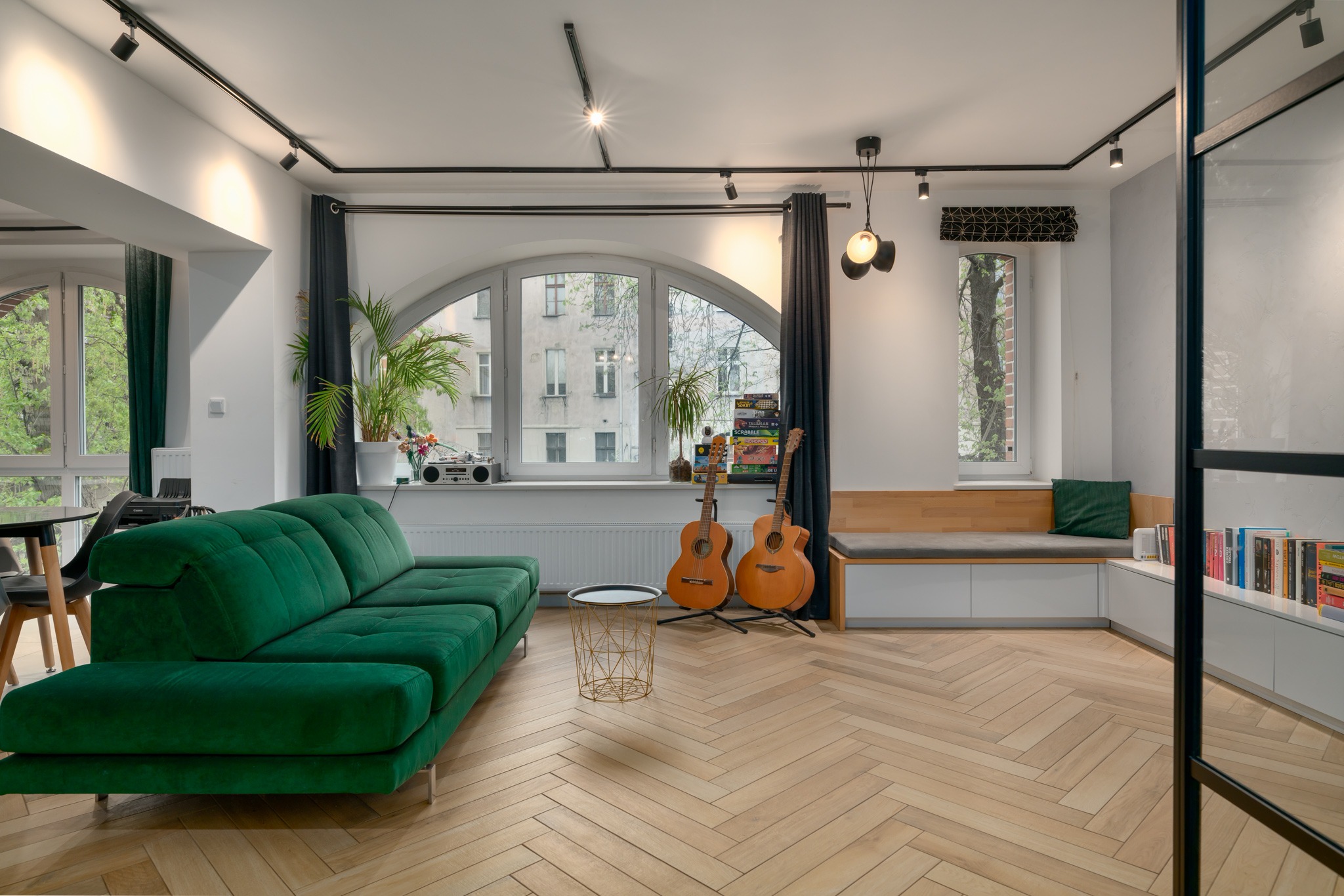 Jasny salon z parkietem w jodełkę, zieloną sofą, dwoma gitarami akustycznymi, roślinami doniczkowymi i oknami z widokiem na budynek mieszkalny.