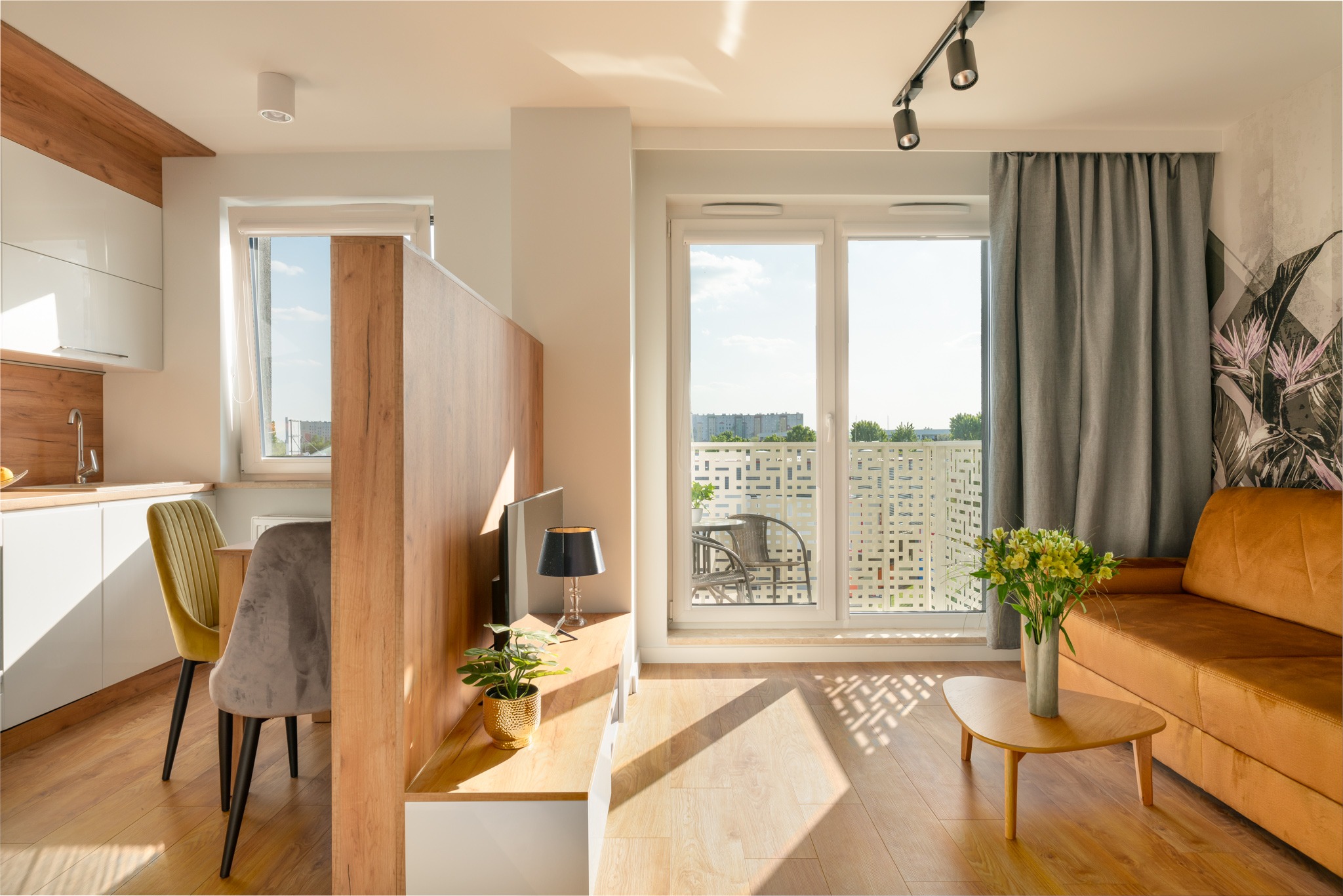 Jasne, nowocześnie urządzone wnętrze mieszkania z aneksem kuchennym, balkonem i częścią wypoczynkową z sofą w kolorze musztardowym oraz bukietem kwiatów na stoliku