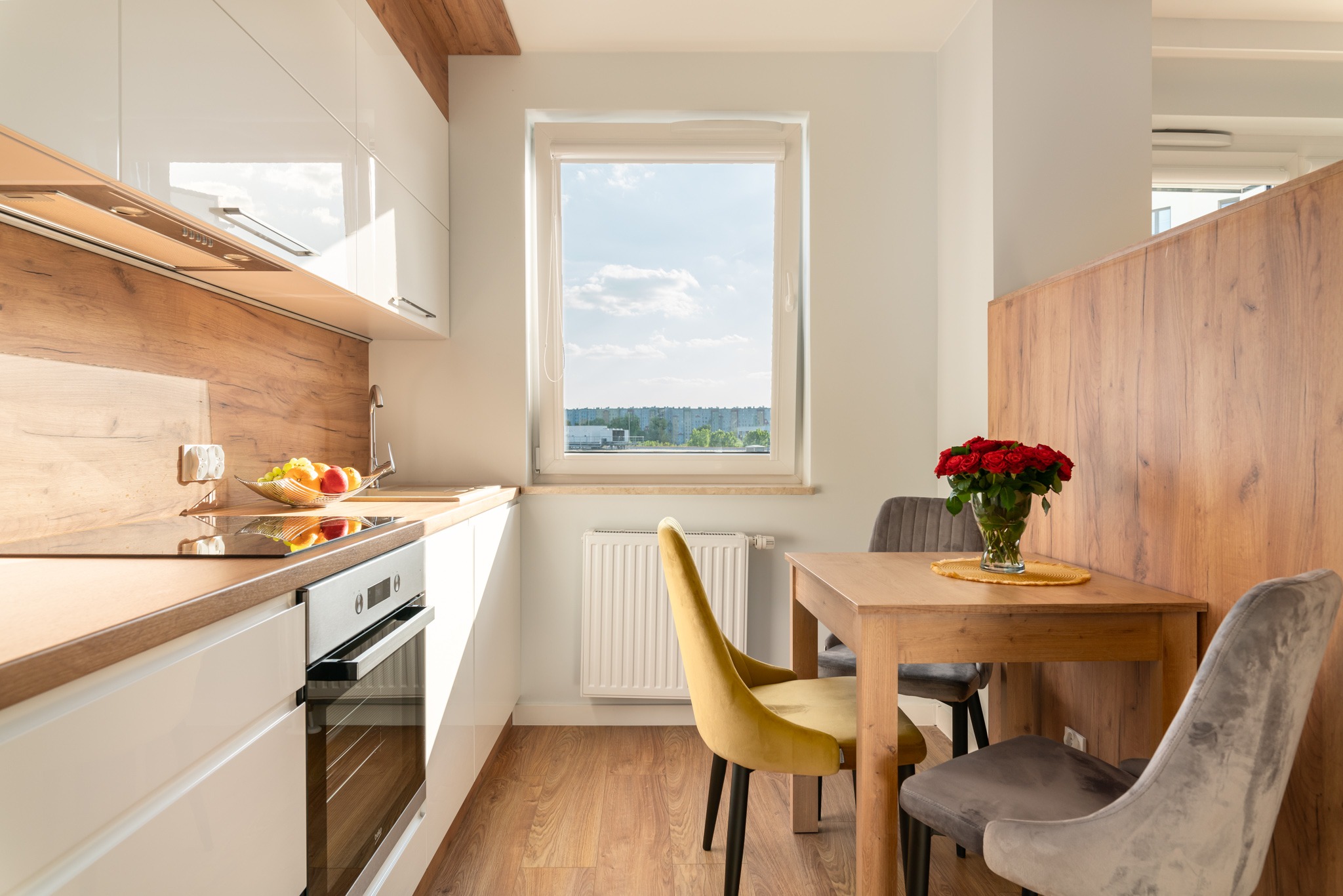 Jasna kuchnia z drewnianymi akcentami, widok na stół z bukietem czerwonych róż, nowoczesne krzesła i okno z widokiem na miasto w tle.