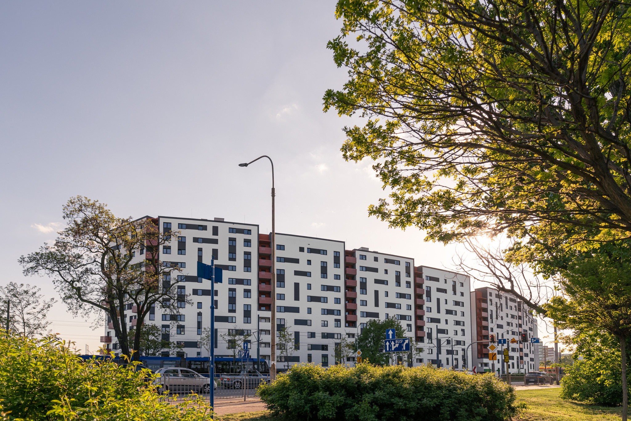 Nowoczesny budynek mieszkalny z białymi elewacjami i czarnymi oknami, widoczny z poziomu ulicy, z zielenią w pierwszym planie i lampą uliczną po lewej stronie.