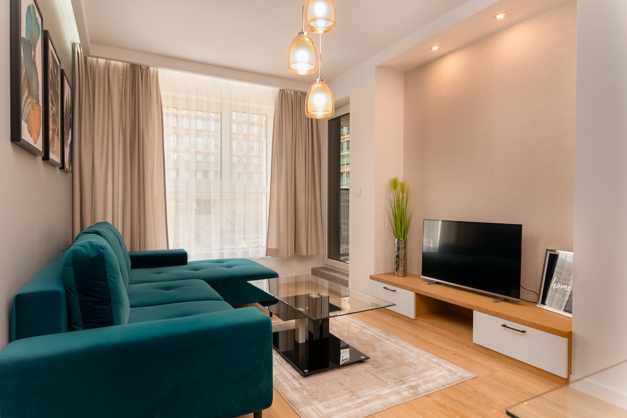 Nowoczesny salon z turkusową sofą narożną, szklanym stolikiem kawowym i drewnianą szafką RTV z telewizorem, ozdobiony beżową tapetą i podświetleniem LED.