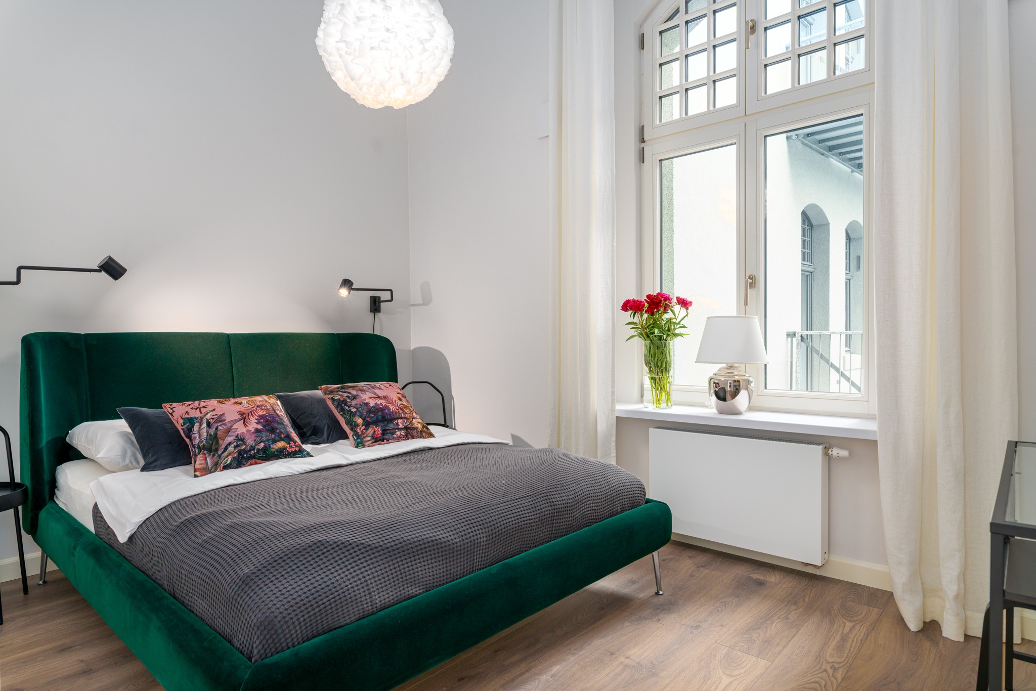Nowoczesna sypialnia z zielonym, welurowym łóżkiem, wzorzystymi poduszkami i drewnianą podłogą, oświetlona lampą wiszącą i lampkami ściennymi, z widokiem na budynek przez okno z białymi zasłonami.