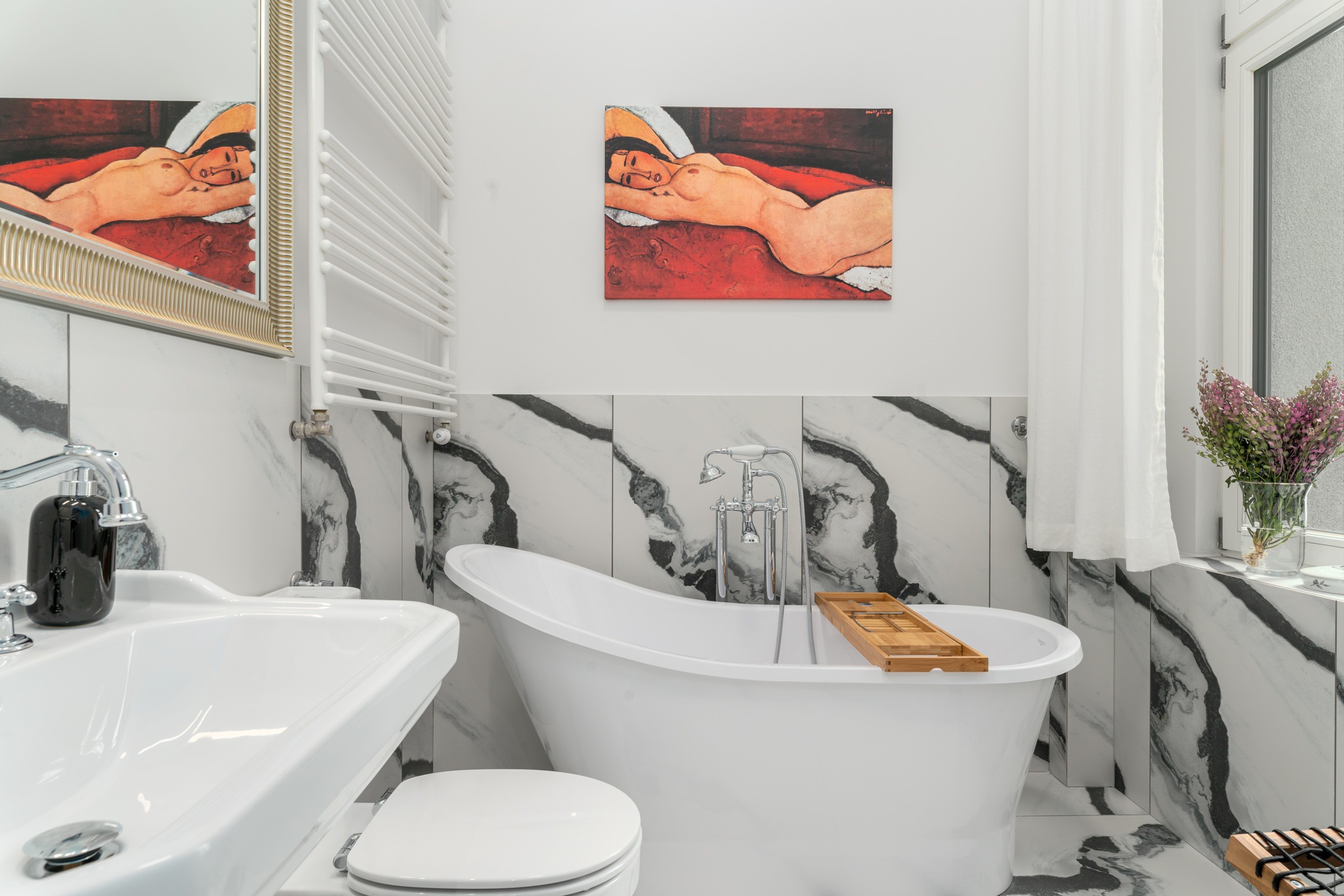 Elegancka łazienka z białą wanną wolnostojącą, marmurowymi płytkami na ścianach i podłodze, obrazem w stylu Modiglianiego, grzejnikiem drabinkowym oraz bukietem kwiatów przy oknie