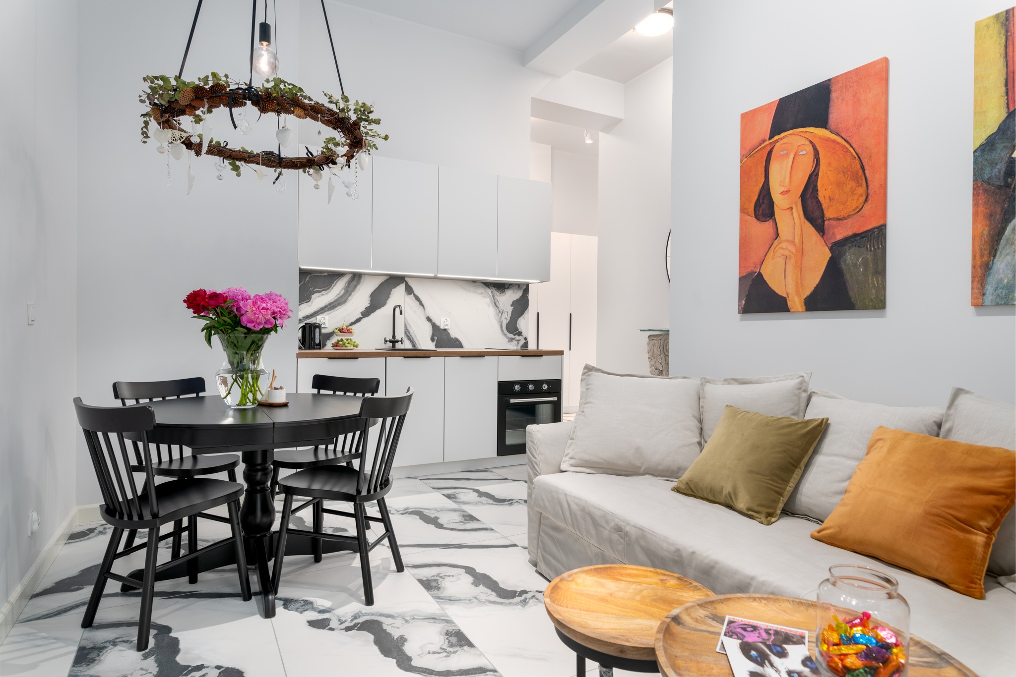 Jasne wnętrze mieszkania z czarnym okrągłym stołem z bukietem różowych piwonii, białą sofą z kolorowymi poduszkami, marmurową podłogą i nowoczesną kuchnią w tle, oświetlone dekoracyjnym żyrandolem...