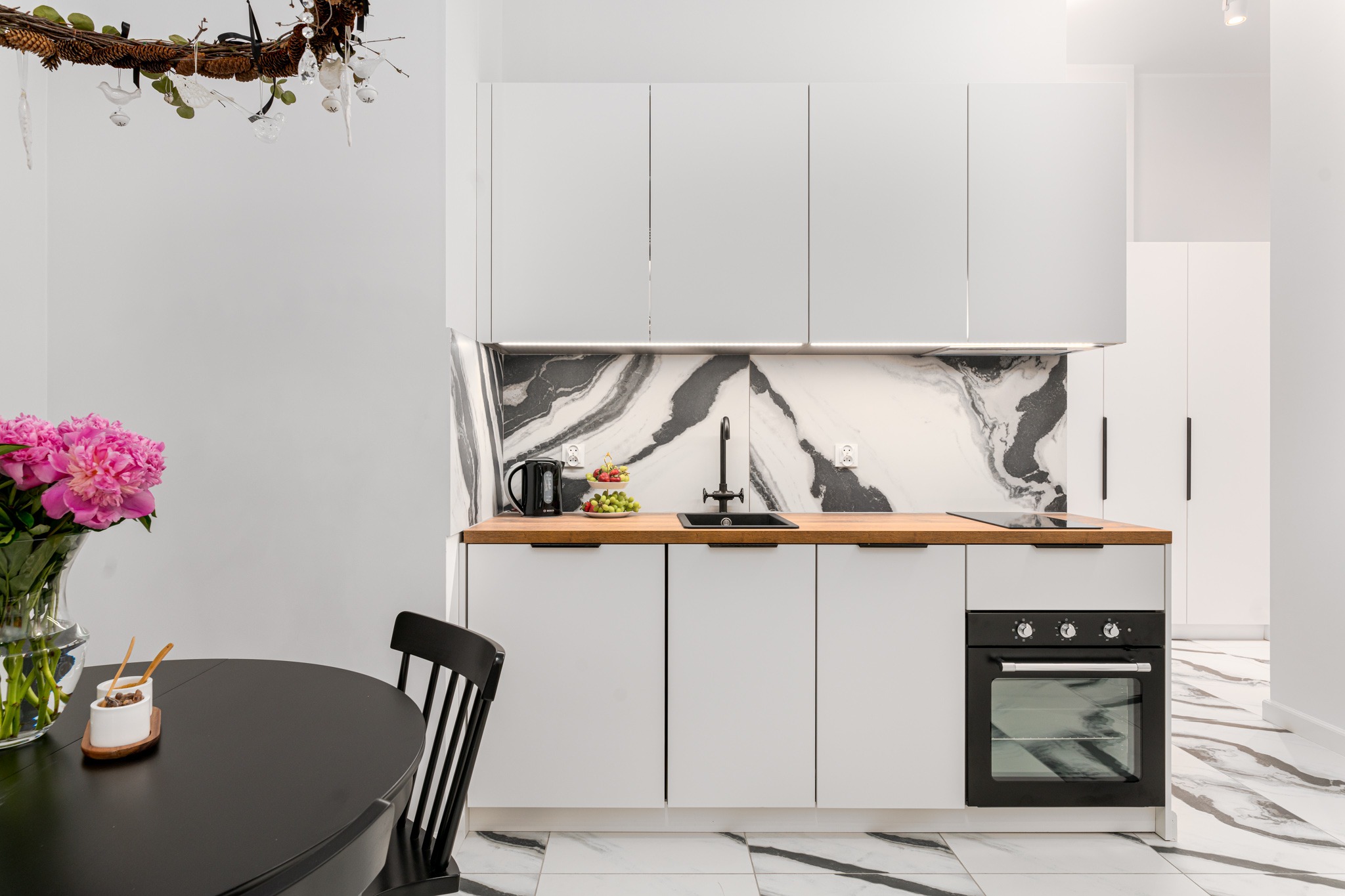 Nowoczesna, jasna kuchnia z białymi szafkami, drewnianym blatem, czarnym zlewem i marmurowym backsplashiem. Na blacie czajnik i owoce. Na podłodze białe płytki z czarnymi żyłami imitującymi marmur.