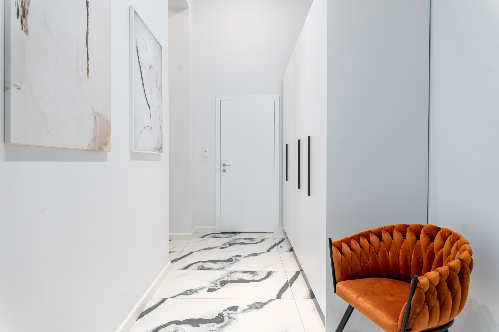 Nowoczesny korytarz z białymi ścianami, marmurową podłogą, minimalistycznymi obrazami i pomarańczowym fotelem z plecionym oparciem.